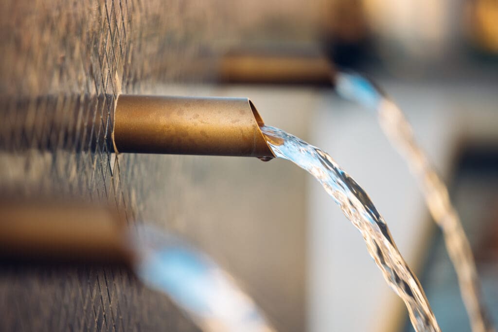 Close-up of water flowing from several brass spouts embedded in a textured wall, creating a cascading fountain effect. The background is softly blurred, emphasizing the smooth, clear streams of water and the reflective surface of the brass spouts.