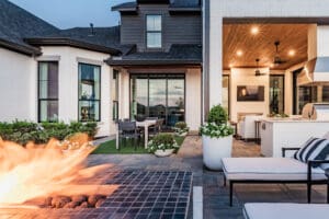Outdoor patio area of a modern house at dusk, featuring a slated fire pit in the foreground, a dining table with chairs on a grassy surface, and a covered lounge area with cushioned seating and a TV. The setting includes lush greenery and contemporary design elements.