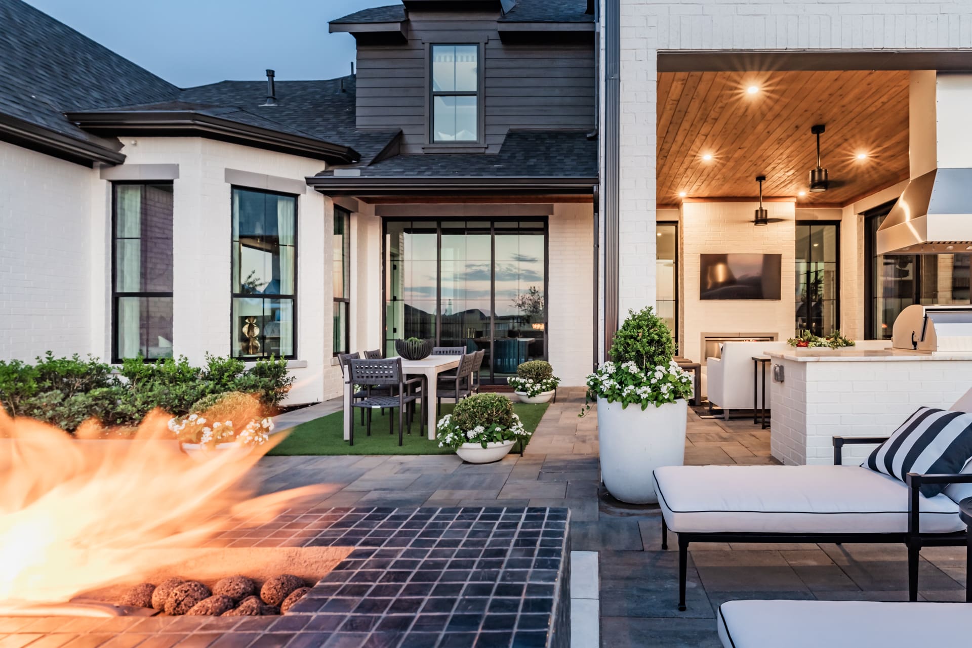 Outdoor patio area of a modern house at dusk, featuring a slated fire pit in the foreground, a dining table with chairs on a grassy surface, and a covered lounge area with cushioned seating and a TV. The setting includes lush greenery and contemporary design elements.