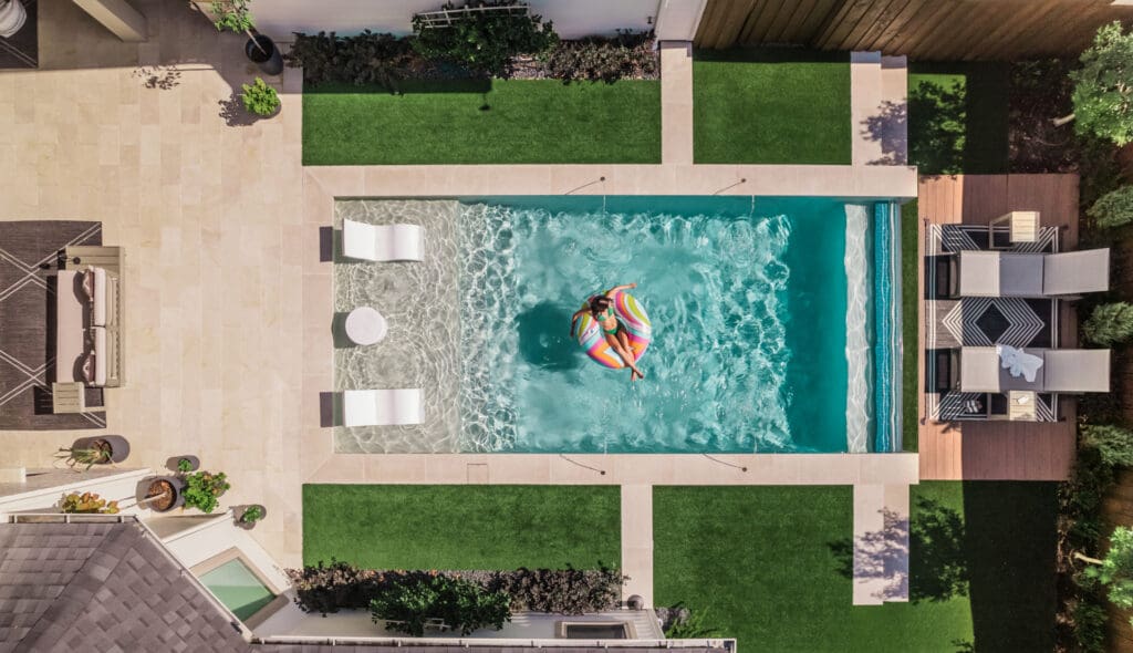 Aerial view of a rectangular swimming pool surrounded by a patio and green lawn. A person is floating on a rainbow-colored inflatable in the pool. There are lounge chairs, a small table, plants, and a seating area with a pergola nearby.