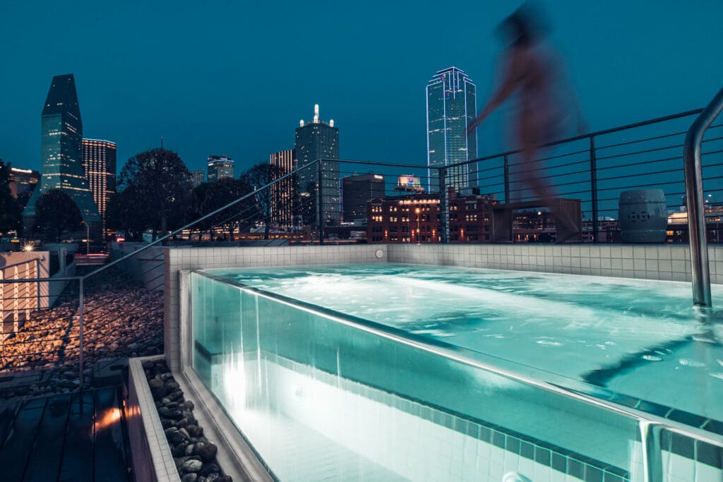 Rooftop pool at night with a cityscape view in the background. A person in motion is walking by the pool's edge. The pool's underwater lights illuminate the water, creating a serene ambiance against the backdrop of tall, lit-up buildings.