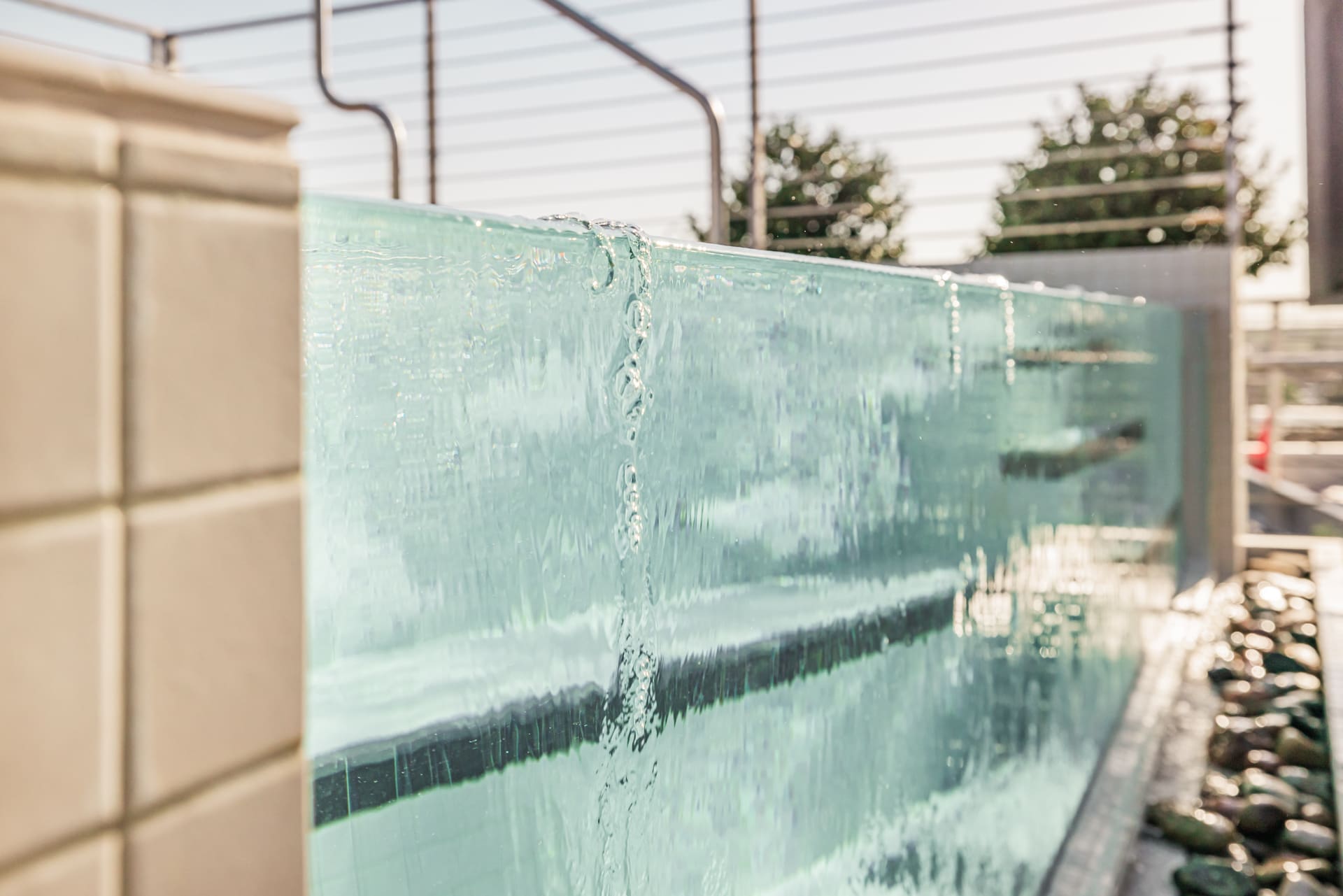 A close-up image of water cascading down the side of a clear glass pool with tiled edges, creating a serene and sparkling effect. In the background, there is a blurred view of trees and metal railings, under a sunny sky.