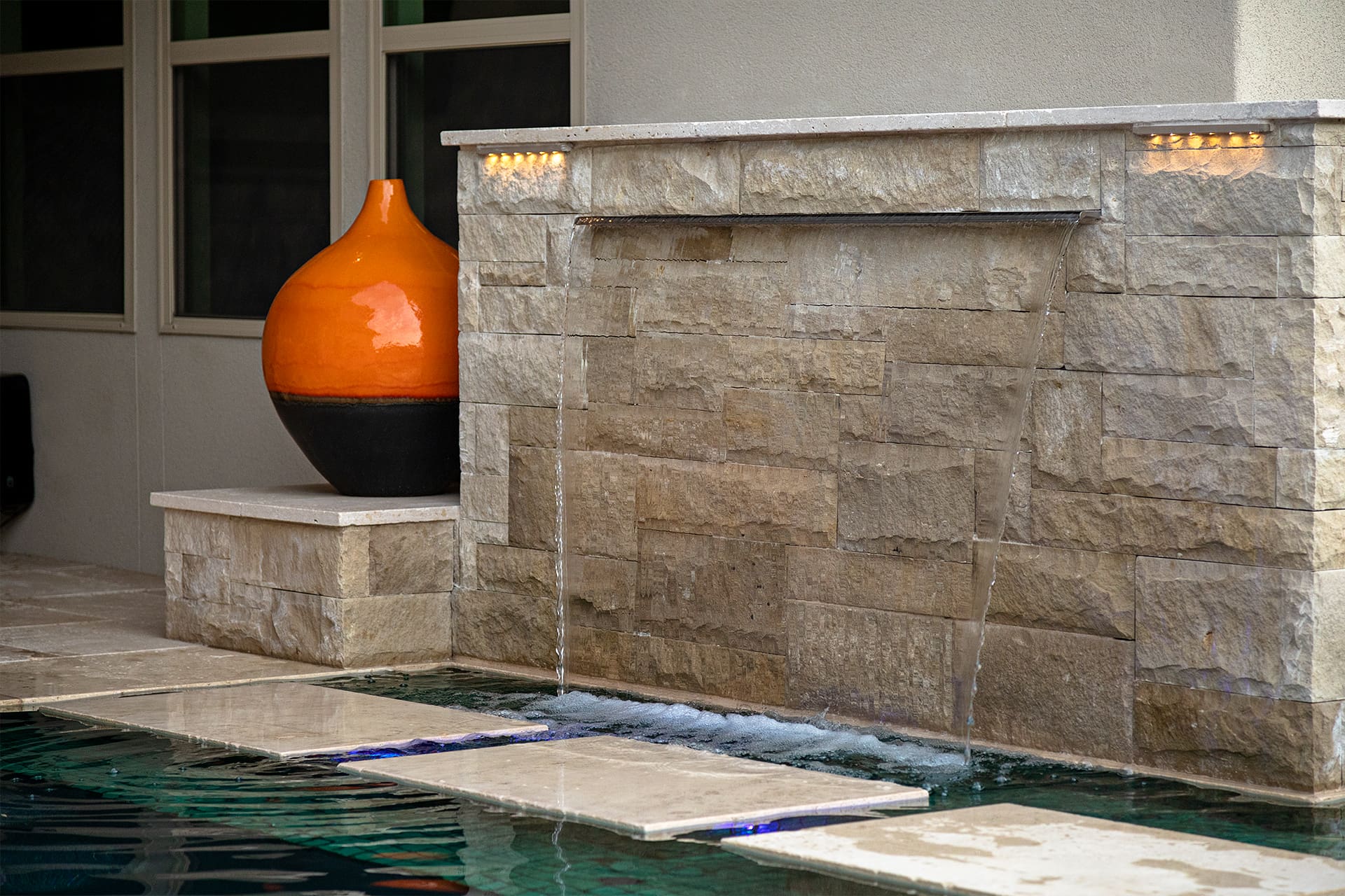 A modern stone water feature with two cascading streams flows into a pool. An orange ceramic vase with a black base sits on a pedestal to the left, adding a pop of color. The setting is serene and contemporary, with clean lines and natural textures.