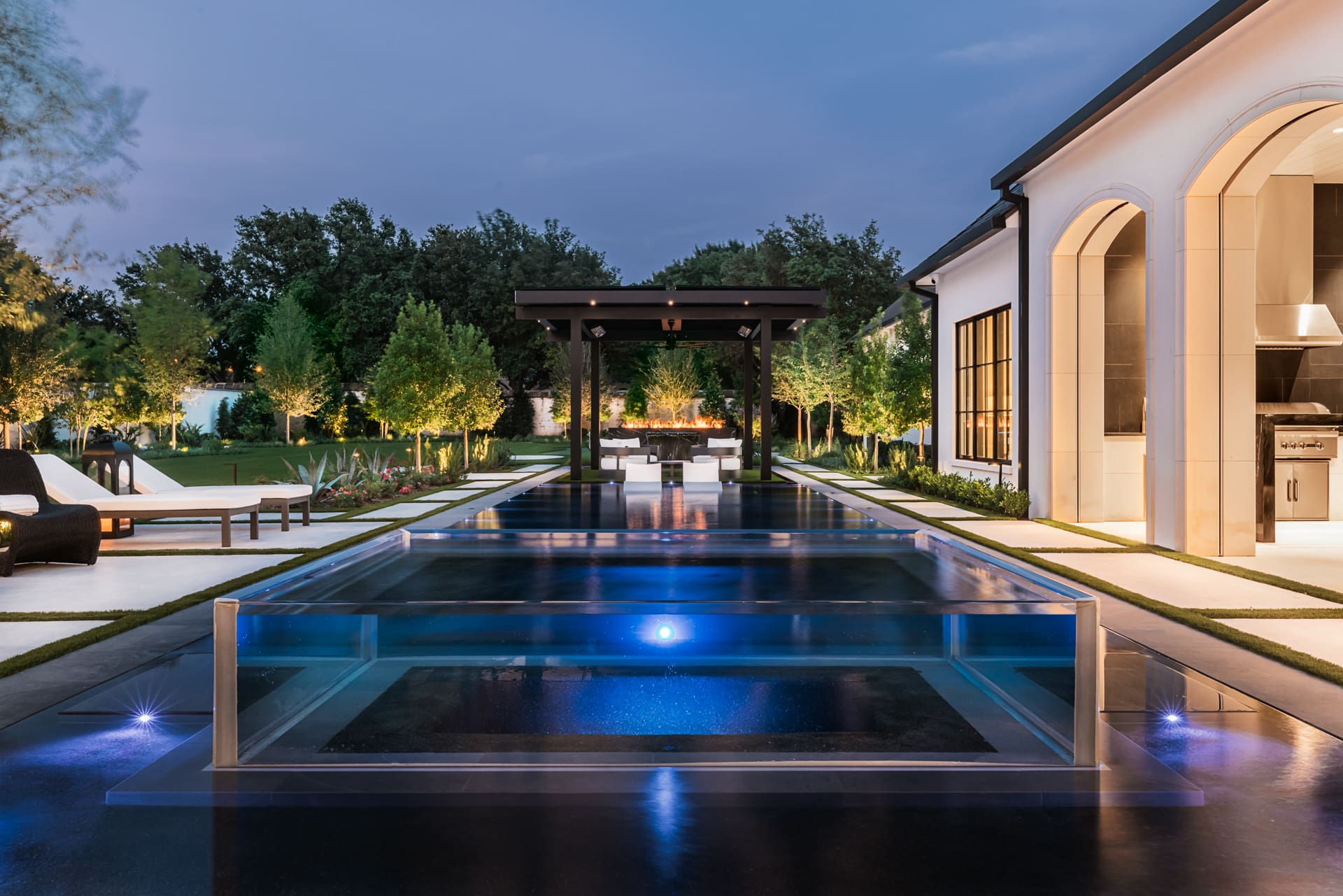A modern, luxurious backyard features a sleek rectangular swimming pool with a glass edge, surrounded by lush greenery and trees. At the far end, a pergola with a built-in fire pit creates a cozy ambiance. A contemporary house with large windows stands to the right.