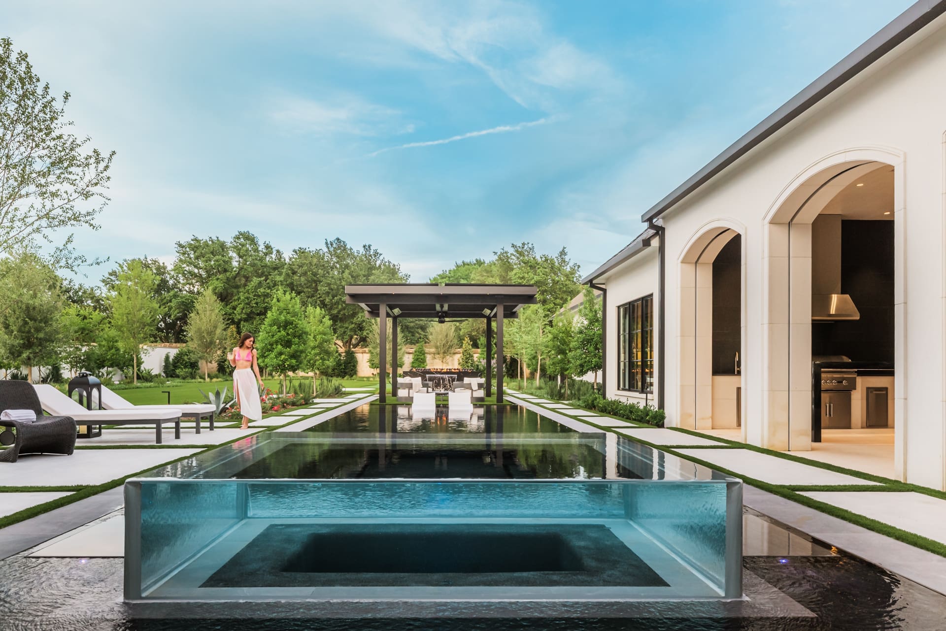 A luxurious backyard features an elegant swimming pool with a transparent edge. A woman in a pink dress stands near the pool, which is surrounded by sleek modern furniture. The space includes an outdoor kitchen and a shaded pergola, all set against lush greenery.