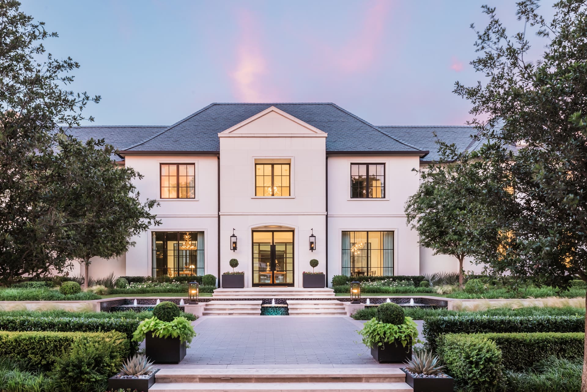 A stately two-story mansion featuring a symmetrical facade with large windows and neatly manicured gardens. Tall trees frame the structure, and steps lead to the entrance, which is accentuated by elegant lantern-style lights. Twilight hues fill the sky.