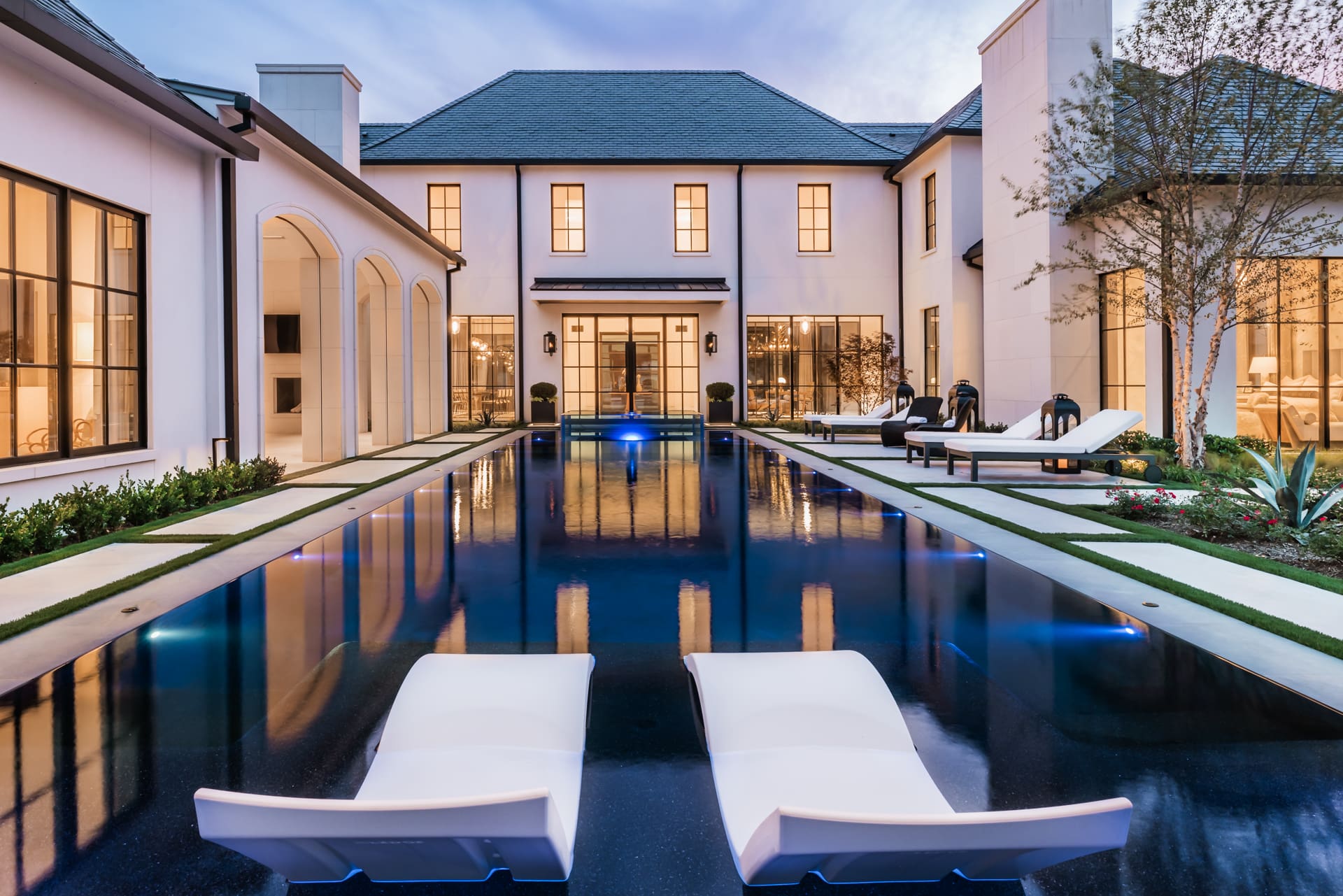 A luxurious modern mansion with a large rectangular pool in the center. The pool is surrounded by neatly trimmed grass and lounge chairs. The house features tall, narrow windows and a mix of modern and classical architectural elements. The sky is at dusk.