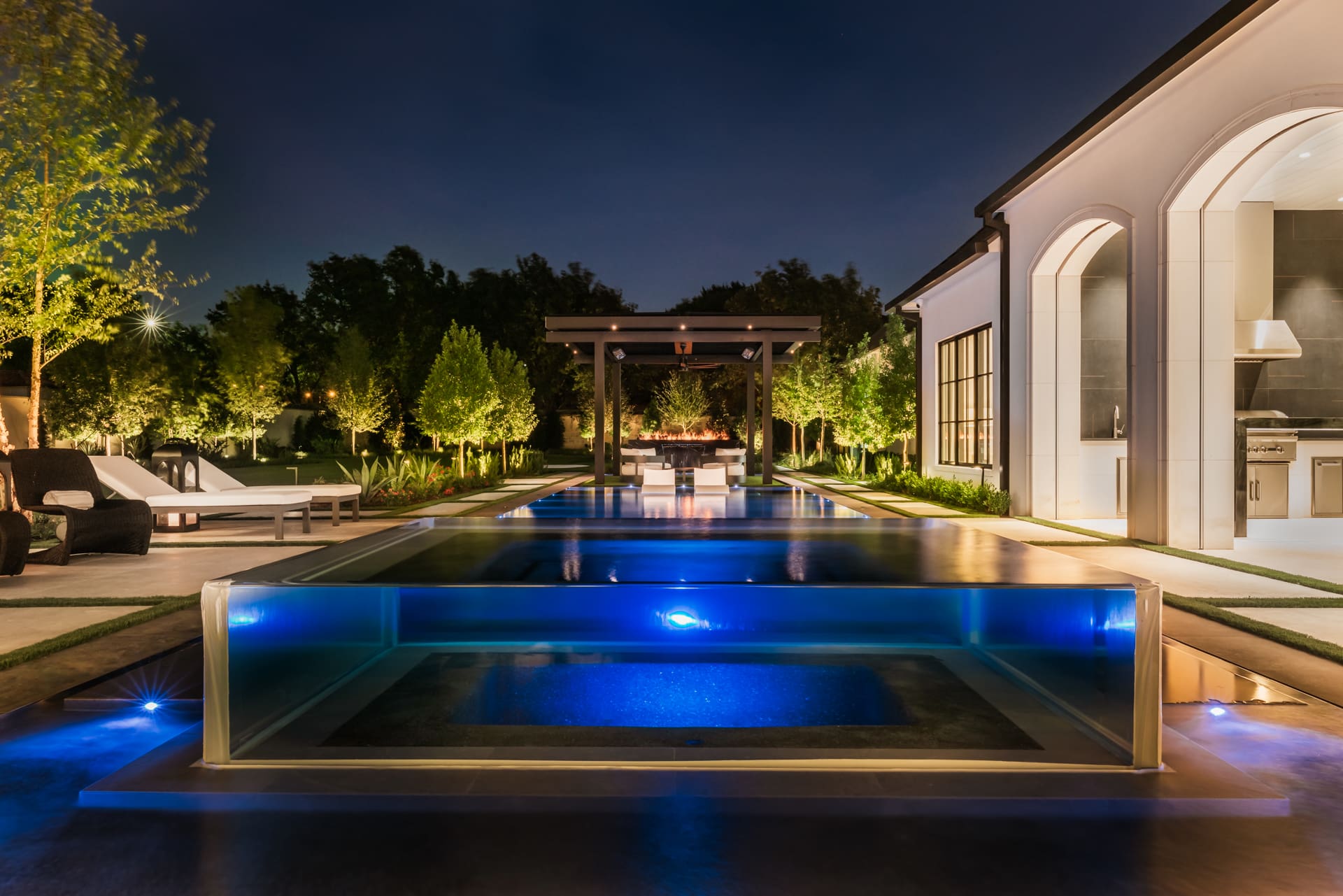 Luxurious modern backyard at night featuring a lit swimming pool with a glass wall, surrounded by greenery and lounge chairs. A pergola with seating is visible in the distance, alongside a well-lit, contemporary outdoor kitchen.