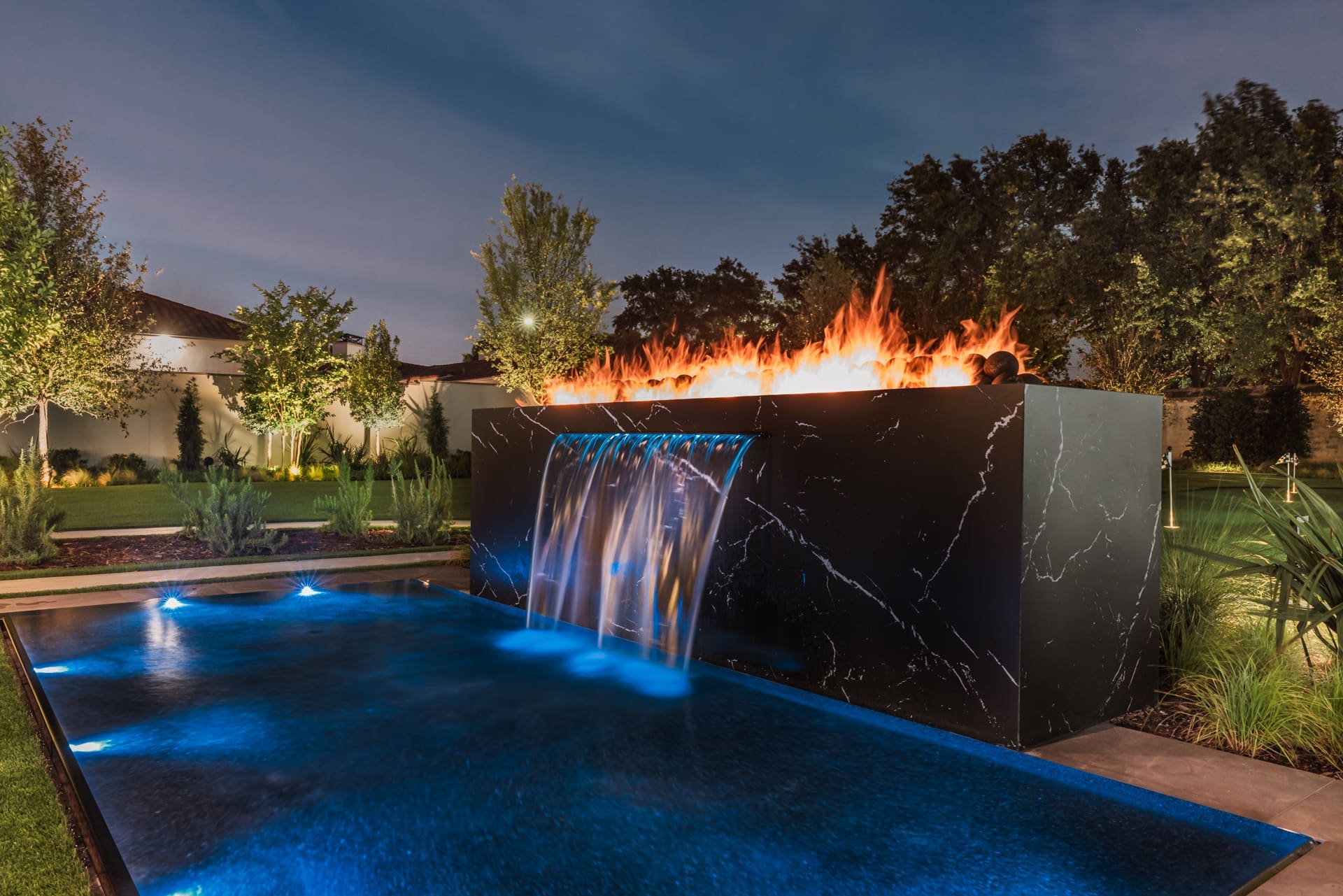A modern outdoor water feature at night, with a sleek black rectangular structure where flames rise from the top. Water cascades from the front into a pool below, which is illuminated with blue lights. Surrounding greenery and a fence are visible in the background.