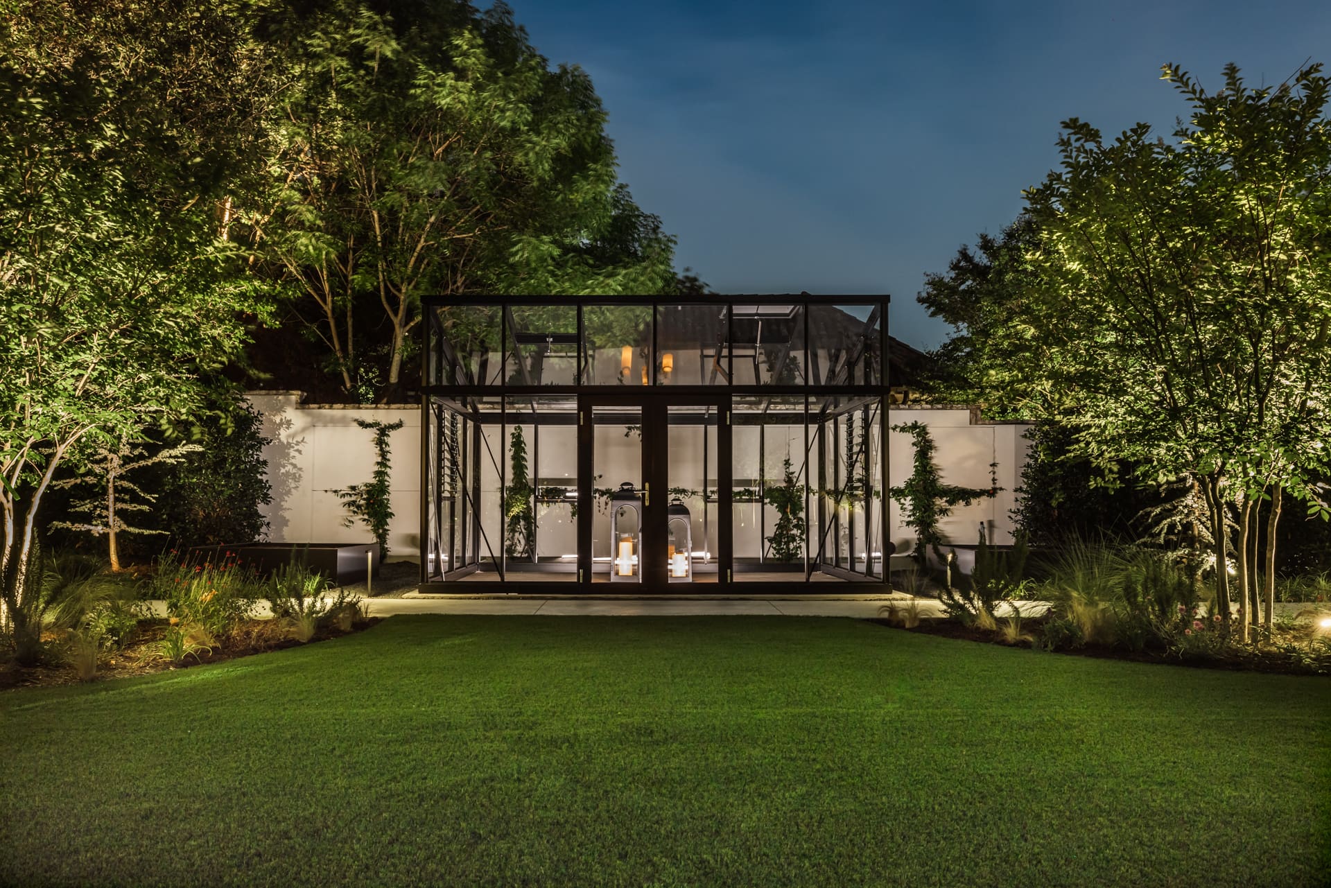 A modern glass and steel structure in a lush garden setting, illuminated at night. Trees and shrubs surround the building, with a neatly manicured lawn in the foreground. Inside, white furniture and decor are visible through the large glass windows.