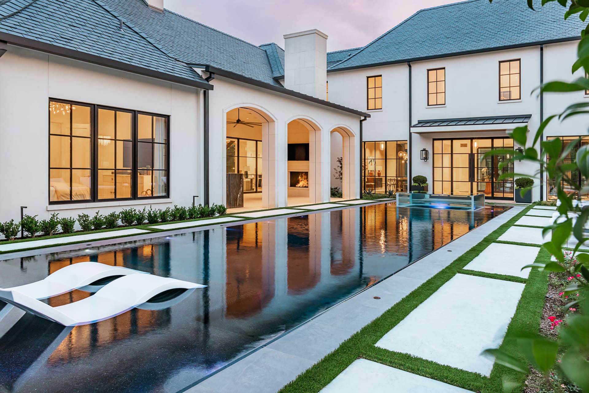A luxurious modern house with large windows and white exteriors features an elegant outdoor pool area. The pool has sleek, white lounge chairs and is surrounded by manicured greenery and stone pavement. The sky is a soft purple and pink, indicating dusk.