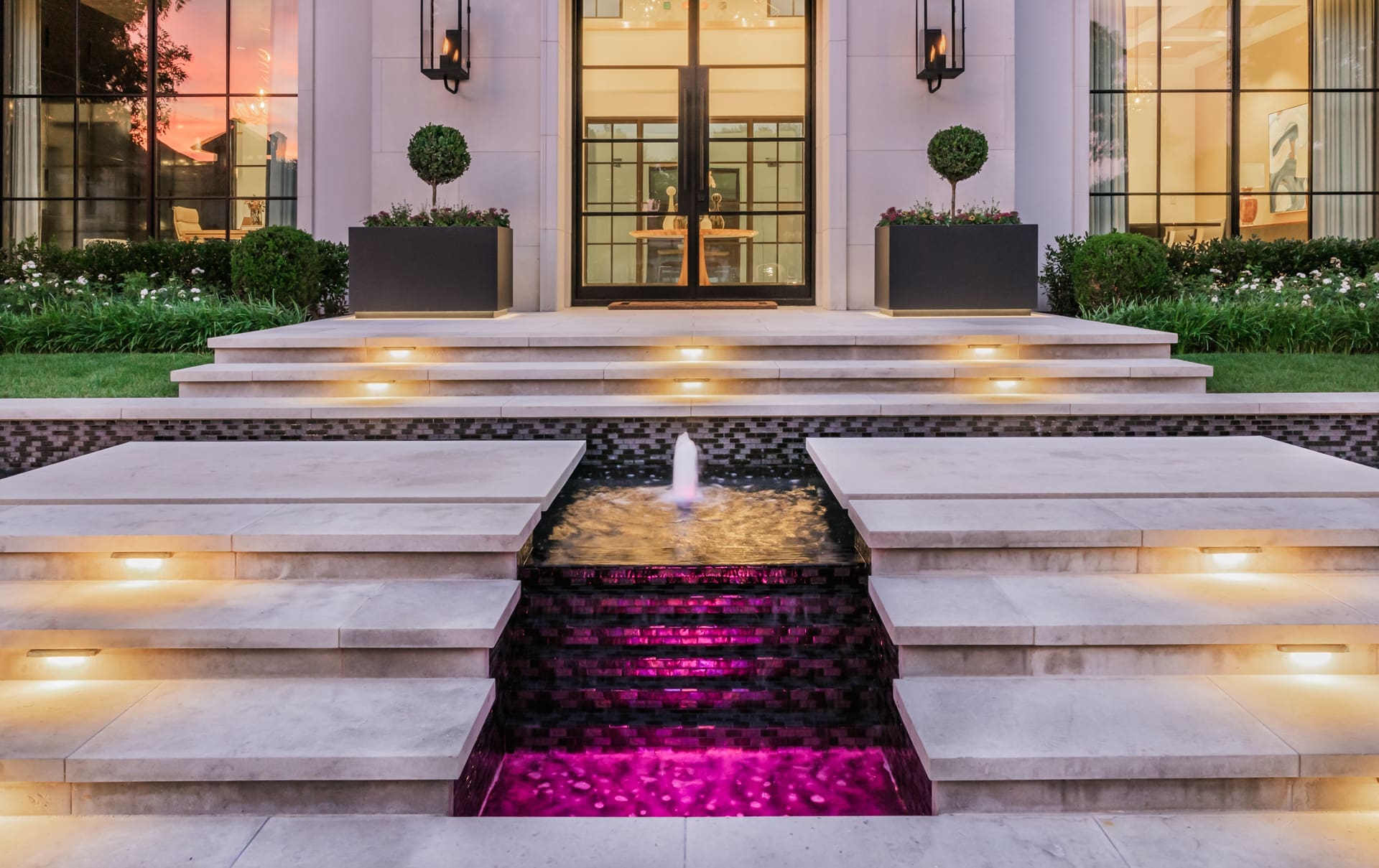 A luxurious modern building entrance with a series of illuminated steps leading up to a glass-door entrance. In front of the steps is a water feature with a central spout and purple lighting. Flanking the entrance are two large planters with neatly trimmed bushes.