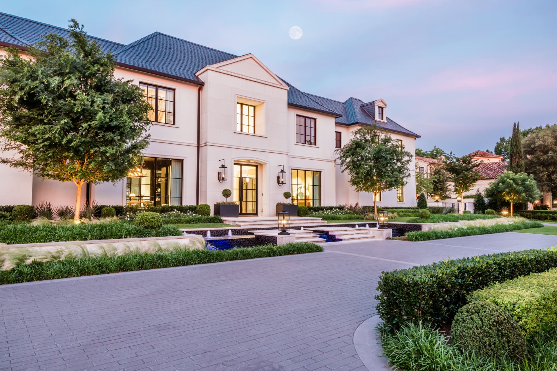 A grand two-story estate sits under a twilight sky with a visible moon. The house has a cream facade and large windows illuminated from within. Elevated by steps, it features a manicured garden with neatly trimmed hedges, trees, and a well-maintained driveway.