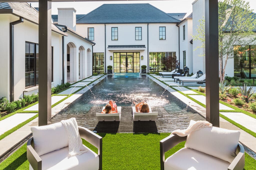 View of a luxurious modern backyard with a large pool, two people relaxing on sun loungers in the water, and contemporary white chairs with cushions in the foreground. The house features large windows and manicured greenery.