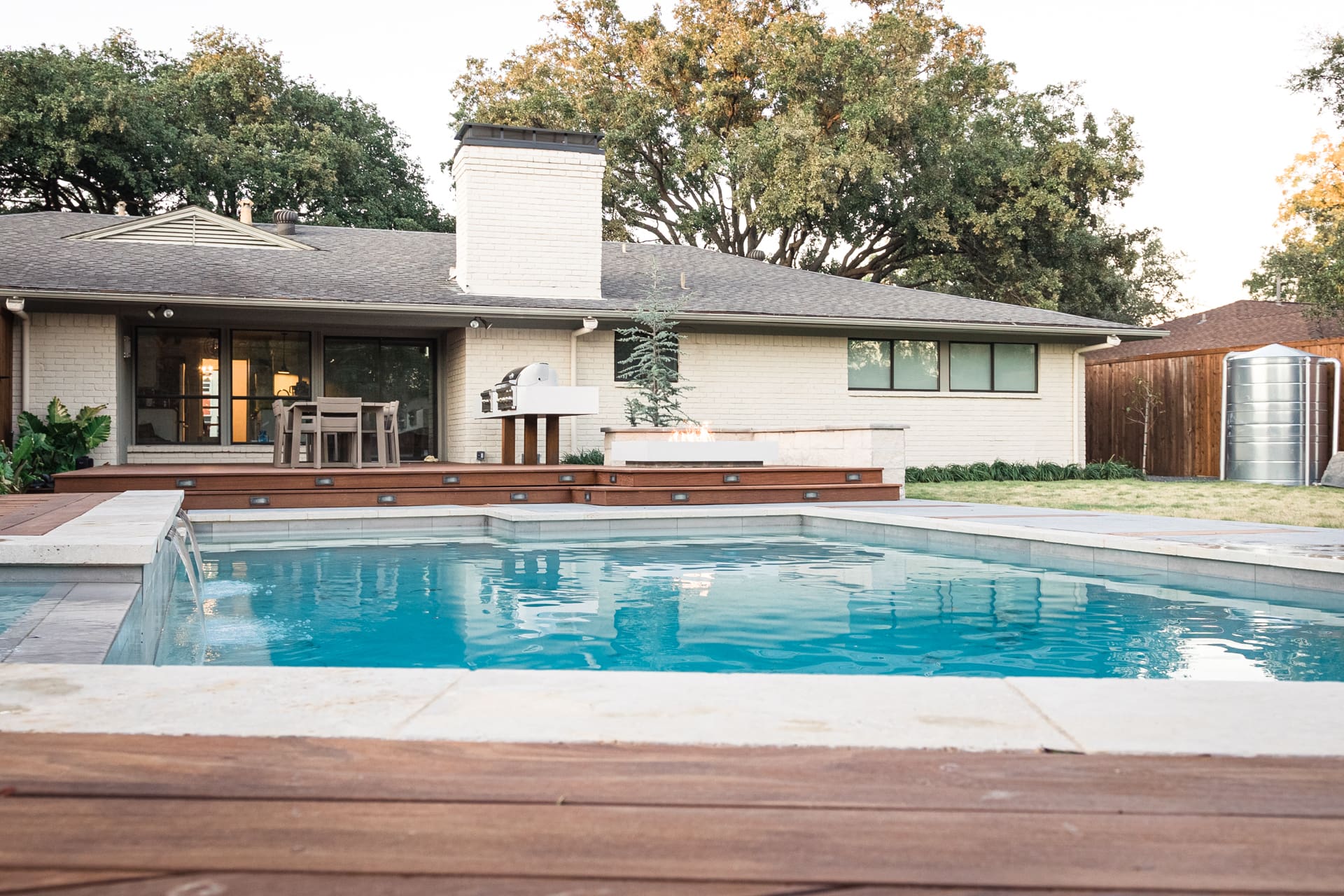 A modern backyard features a rectangular swimming pool with clear blue water. Surrounding the pool is a wooden deck with outdoor furniture and a grill. In the background is a single-story house with large windows and a chimney, bordered by trees and a fenced garden.