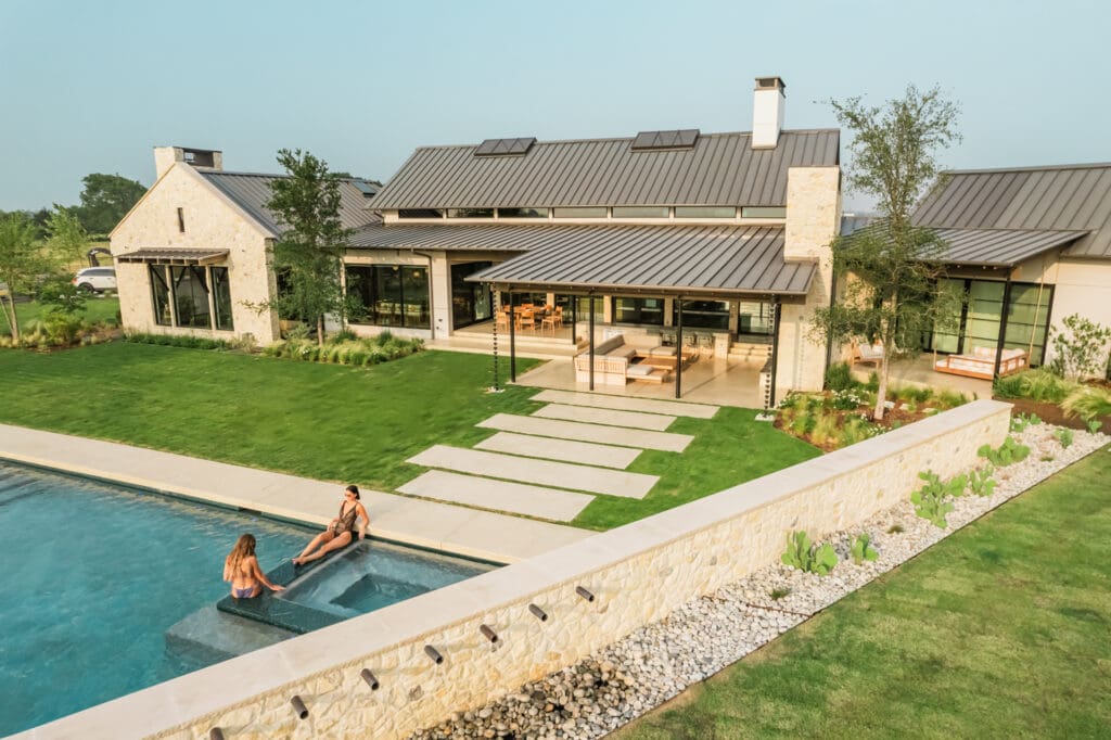 A modern house with a stone facade and metal roof features a large backyard. There is a pool and patio area, where two people are sitting by the water. The well-manicured lawn and pathway add to the serene setting. The house has large windows and multiple trees around.