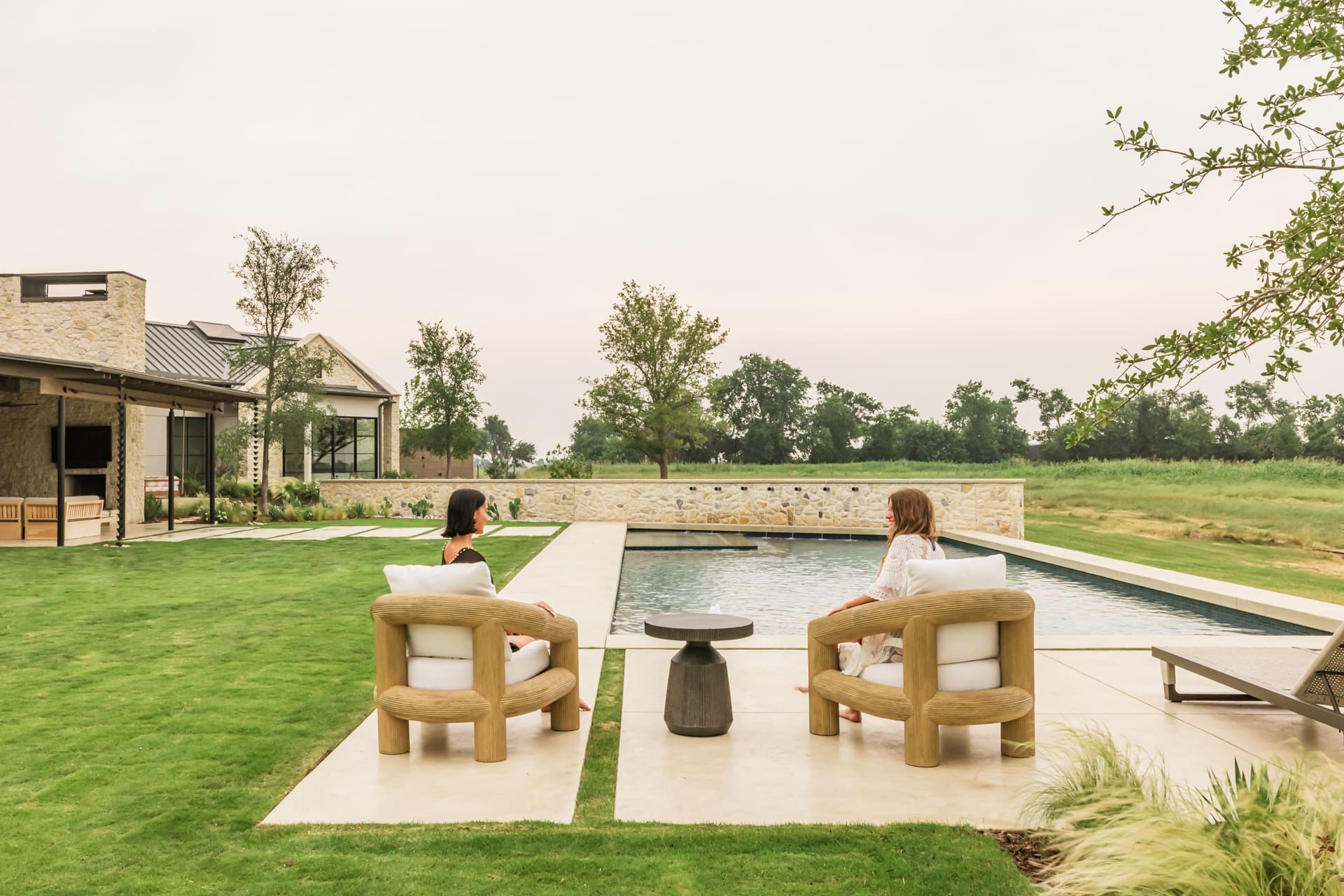 Two people sit on wooden chairs by a modern rectangular pool, overlooking a lush green lawn and distant trees. The scene is serene, with a cloudy sky and a modern house visible in the background.