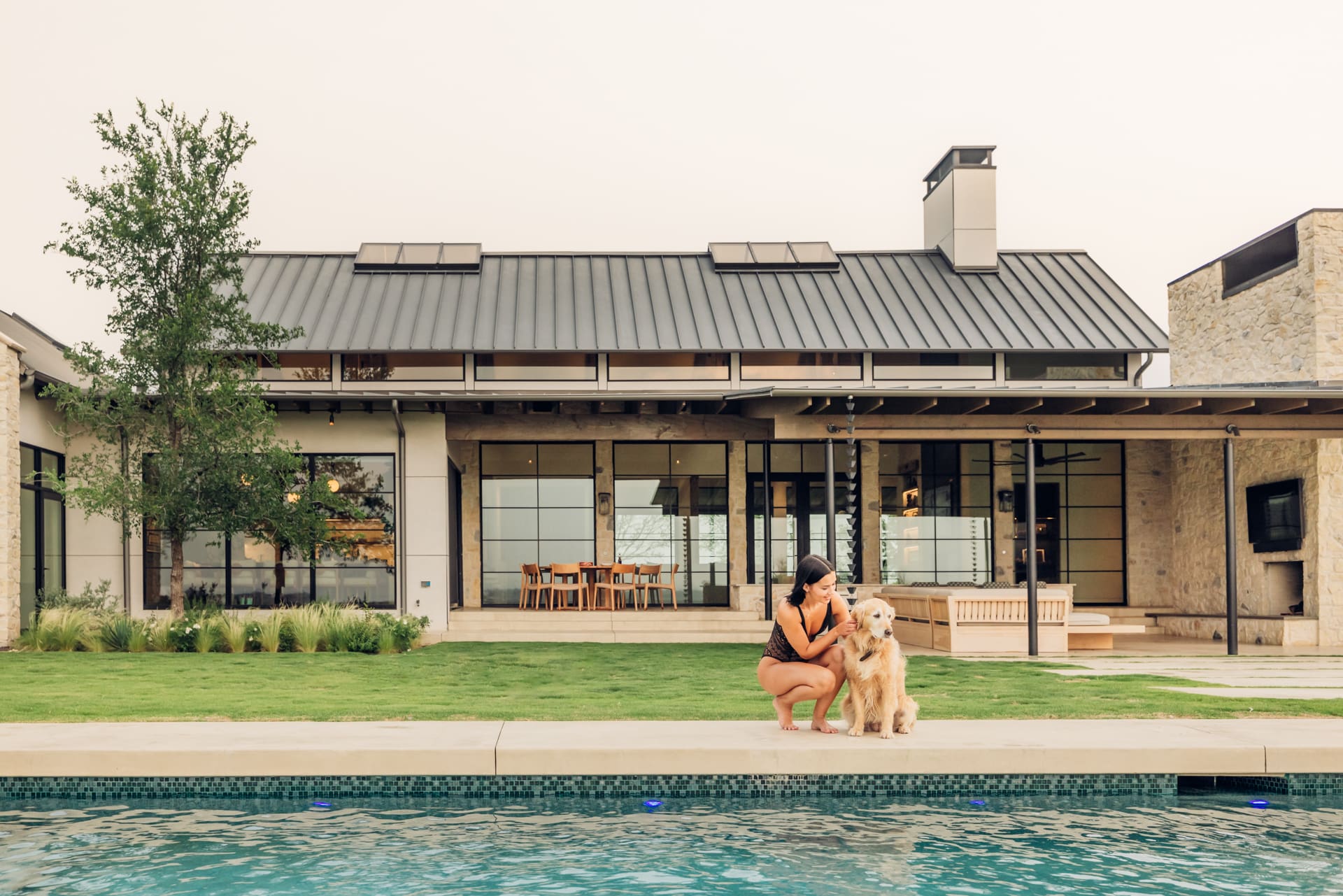 A person kneels beside a golden retriever on the edge of a pool in front of a modern house with large windows and a metal roof. There's a patio with outdoor seating, and the surrounding lawn is well-kept.