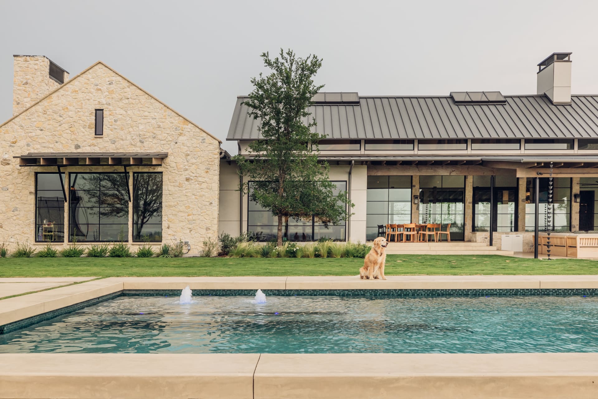 A golden retriever sits beside a rectangular pool with fountains, in front of a modern house featuring stone and metal architecture. The home has large windows and a covered patio area, surrounded by a grassy lawn and trees.
