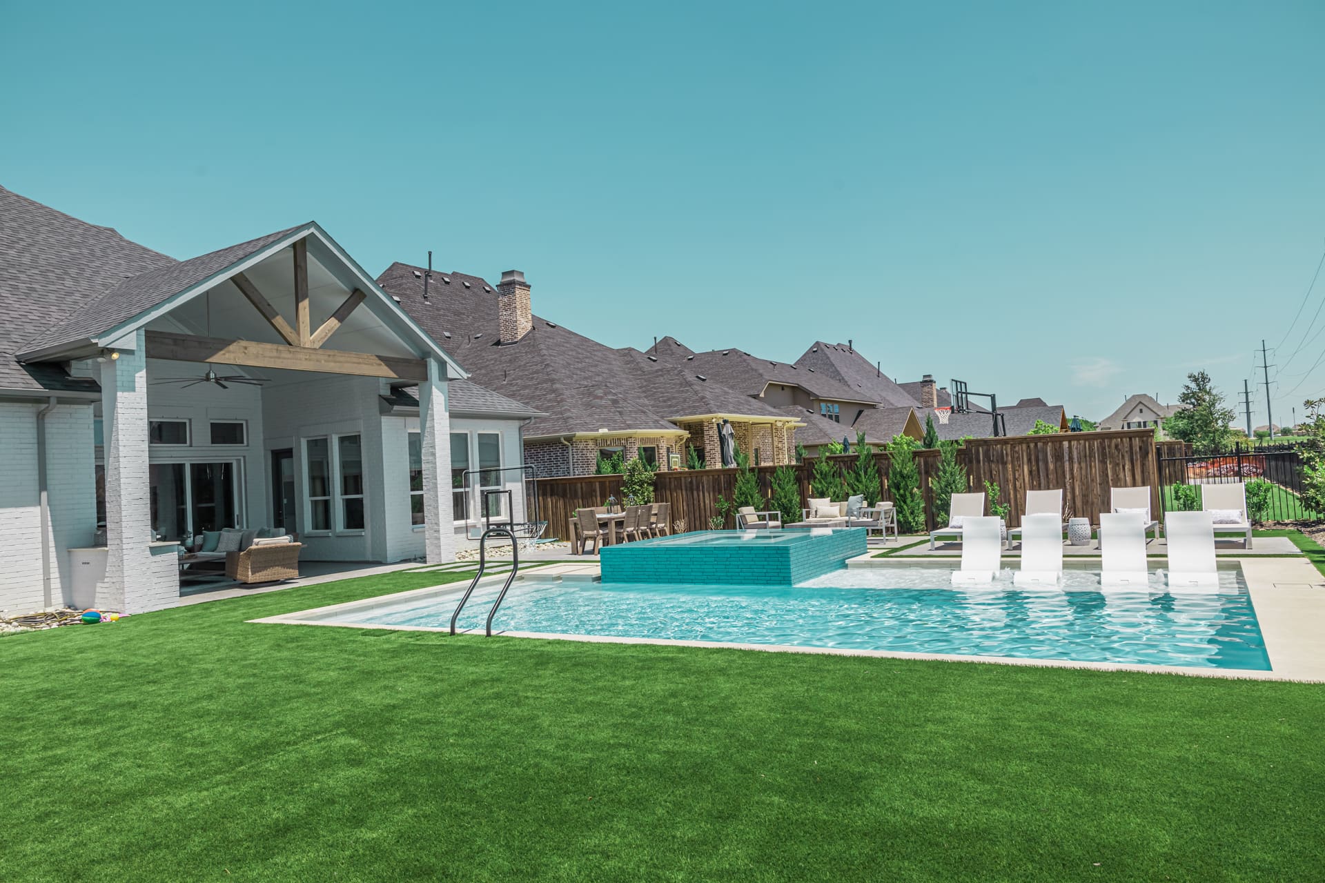 A backyard with a modern swimming pool featuring clear blue water, adjacent to a white house with large windows. The pool has a built-in hot tub and sun loungers on a shallow shelf. The backyard is surrounded by a wooden fence and has well-maintained green grass.