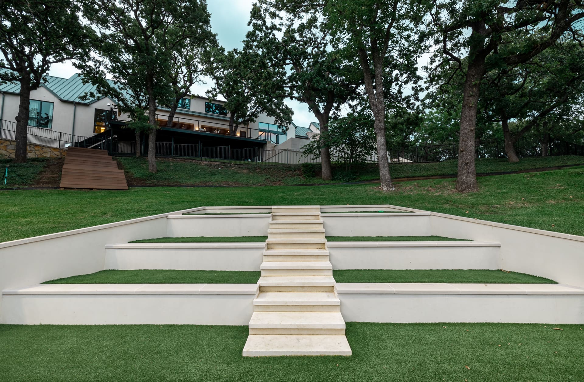 A green lawn featuring a wide staircase with light-colored steps running up the middle, bordered by tiered grass terraces. In the background, there is a modern building atop a slope, with large trees lining the area. The sky is overcast.
