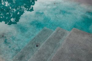 A serene, clear pool with aqua blue water and textured concrete steps leading into it. The water reflects trees and greenery from the surroundings, creating a calm and tranquil atmosphere.