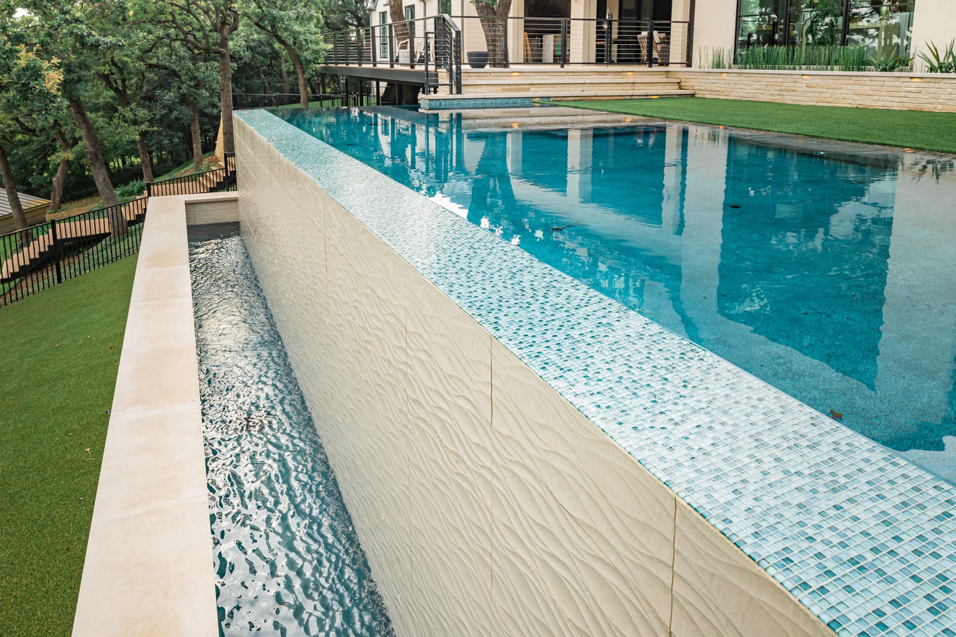A beautiful outdoor infinity pool with clear blue water, elevated above the surrounding landscape. The pool has a cascading water feature along one side, flowing into a narrow trough below. The area is surrounded by lush greenery and modern architecture.