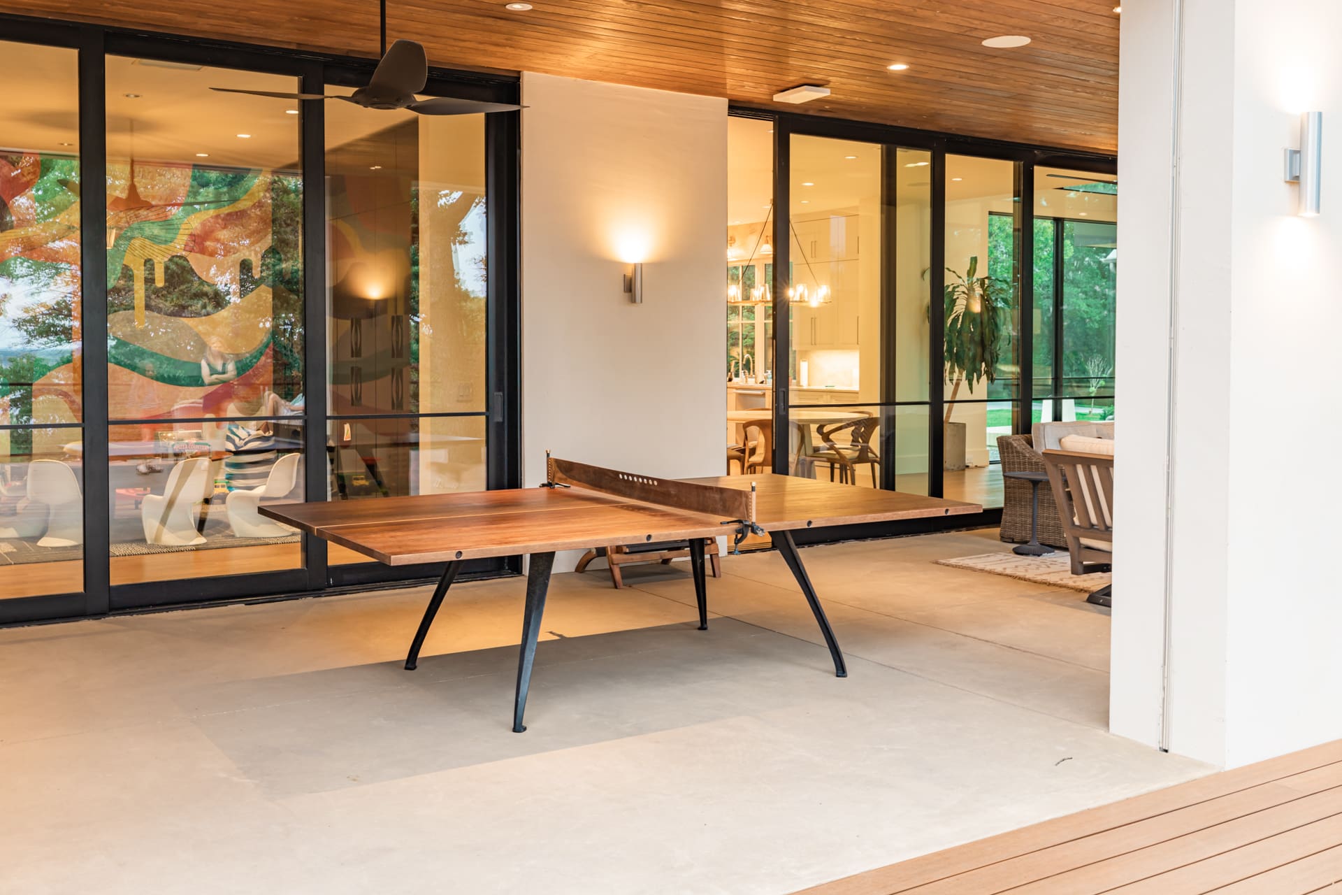 A modern outdoor patio features a wooden ping pong table with sleek black legs. Behind the table, large glass doors reveal an interior art piece and parts of a contemporary living space adorned with plants and warm lighting. The ceiling is wood-paneled.