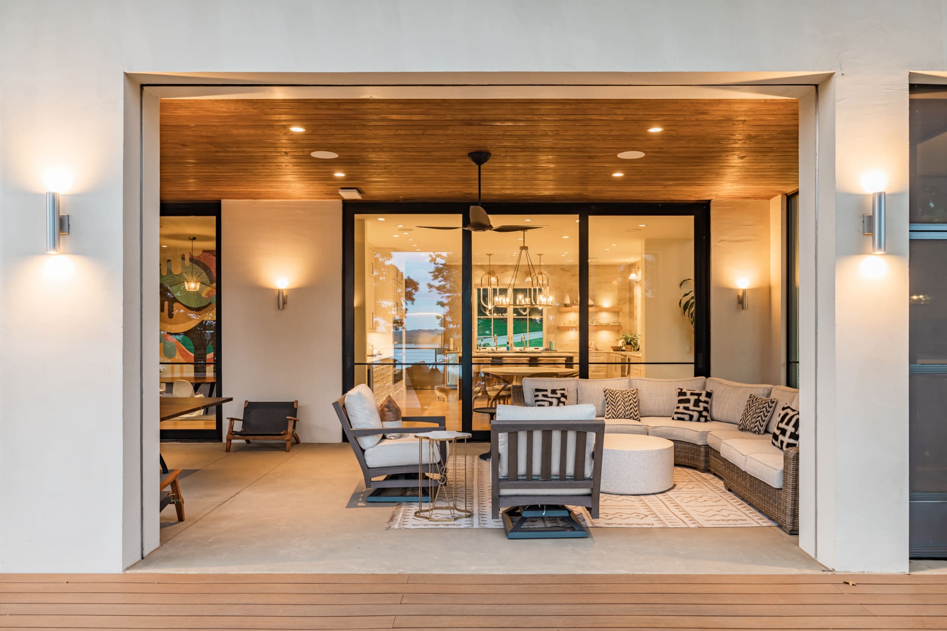A modern, open-concept outdoor living space featuring a comfortable sectional sofa, two armchairs, a round coffee table, and soft ambient lighting. The area opens into a contemporary kitchen and dining room with sleek furnishings visible through large glass sliding doors.