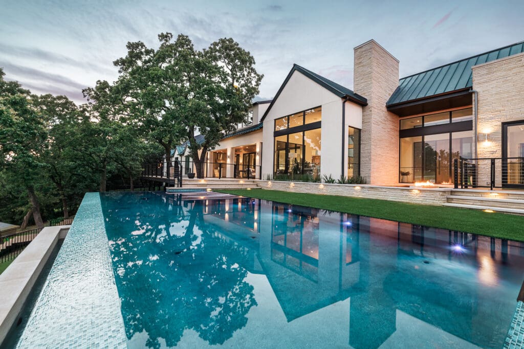 A luxurious modern house with large glass windows sits adjacent to a pristine infinity pool that reflects the evening sky. The surrounding area is adorned with greenery and trees, creating a serene and elegant atmosphere. Lights illuminate the steps leading to the pool.