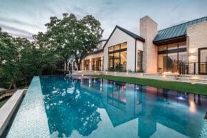 A luxurious modern house with large glass windows sits adjacent to a pristine infinity pool that reflects the evening sky. The surrounding area is adorned with greenery and trees, creating a serene and elegant atmosphere. Lights illuminate the steps leading to the pool.