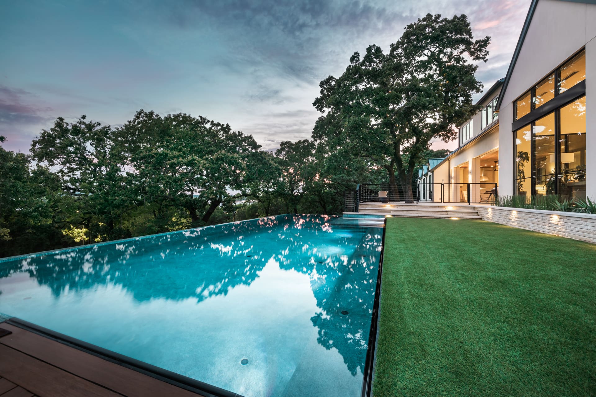 A modern house is visible in the background with large glass windows. An infinity pool reflects the sky, surrounded by trees. The pool area is lined with lighted steps and a well-maintained lawn. The sun is setting, casting a serene ambiance over the scene.