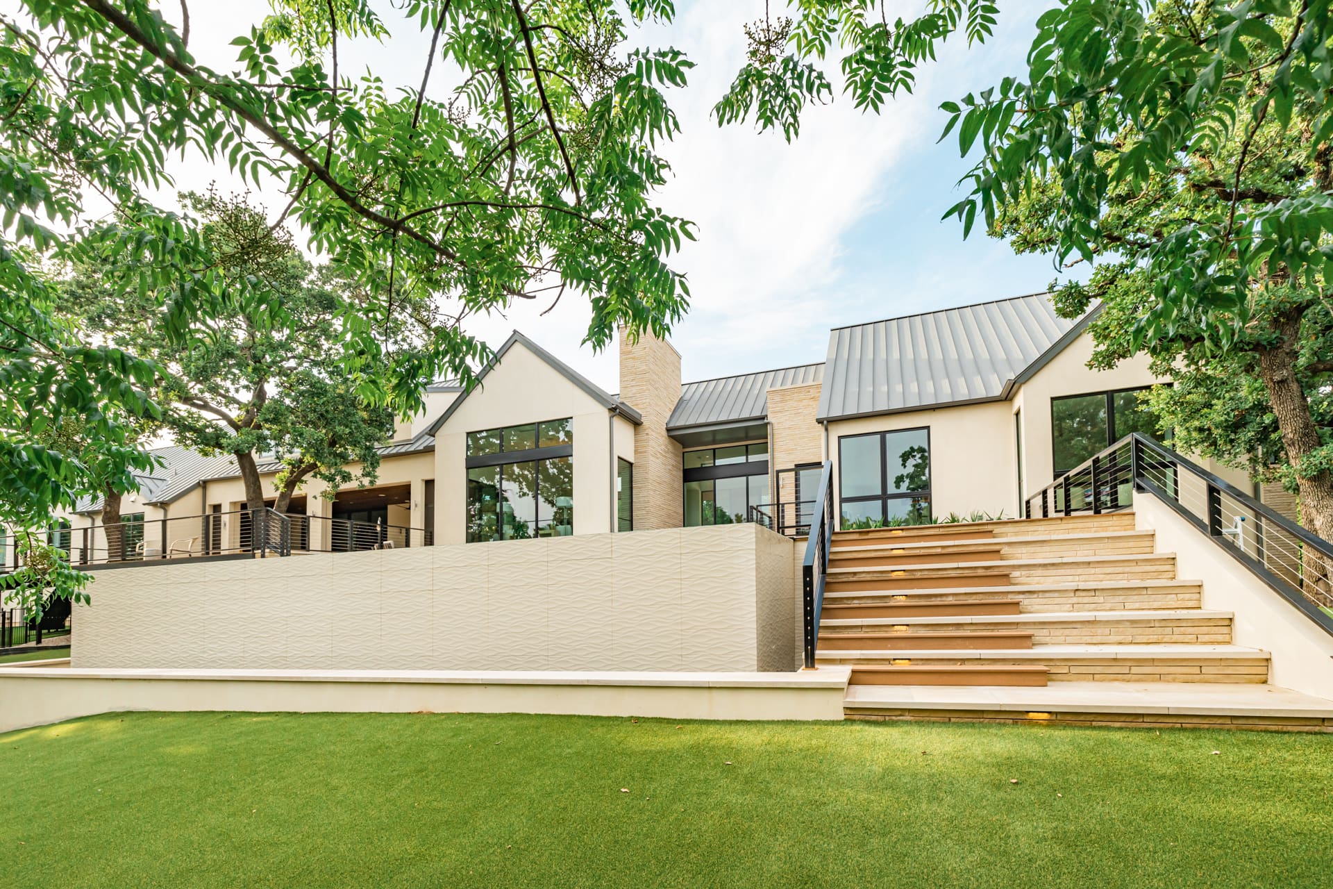 A modern, two-story white house with large glass windows, black frames, and gabled roofs. The home features a broad, ascending staircase that leads to a spacious patio. Tall trees and a well-manicured green lawn surround the property under a bright sky.