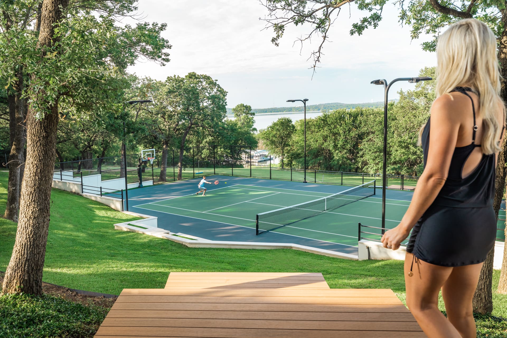 A woman in black workout attire stands on a wooden deck overlooking a green, outdoor tennis court. Another person is playing tennis on the court surrounded by trees, with a glimpse of a lake in the background. The scene is sunny and serene.