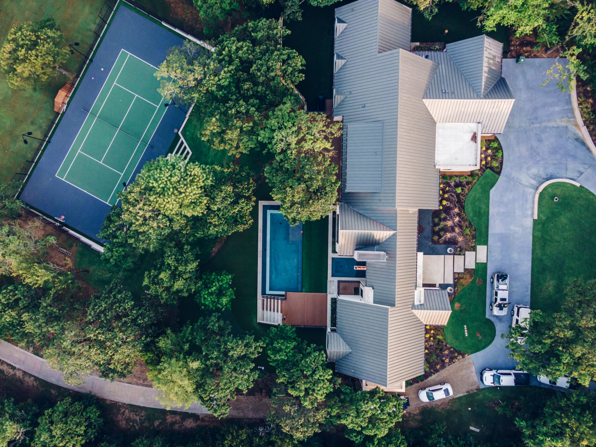 Aerial view of a large modern house surrounded by trees. The property includes a swimming pool, a tennis court, and a driveway with parked cars. The house has a multi-level roof design, and various outdoor spaces are visible.