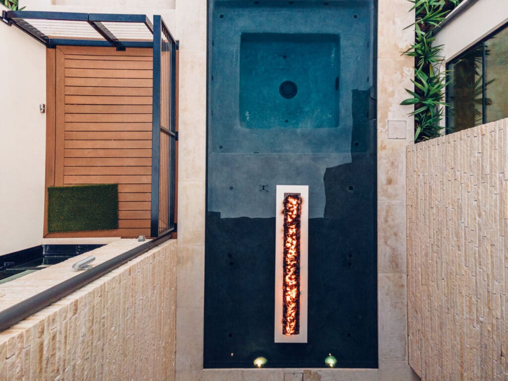 A top-down view of a modern, narrow rectangular swimming pool with a built-in hot tub. The pool features a unique rectangular fire pit in the center, surrounded by tall stone walls and greenery along the edges.