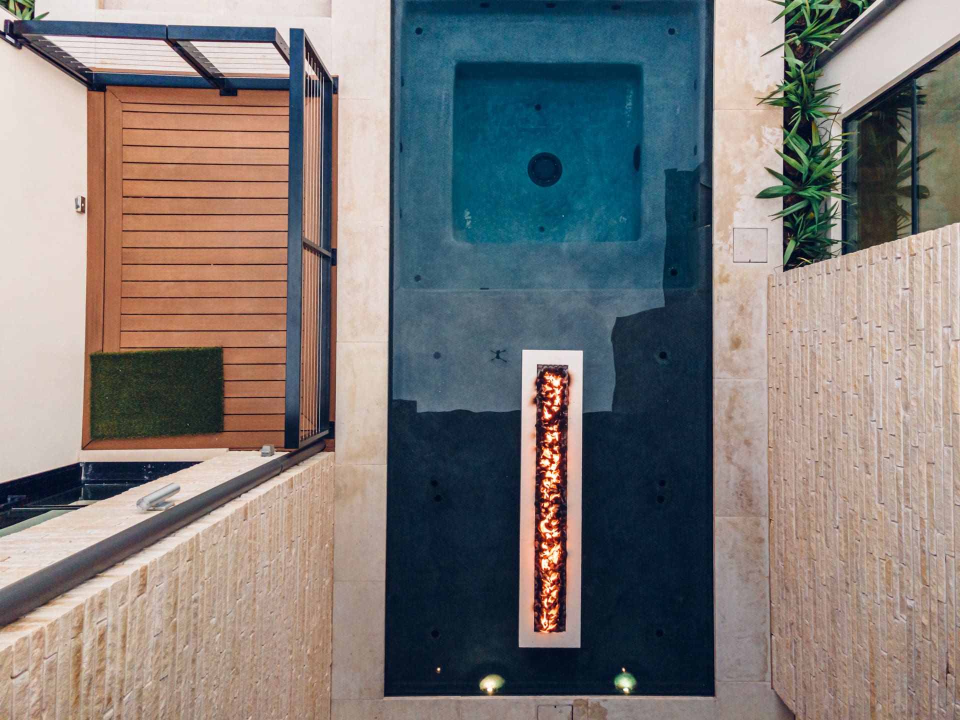 A top-down view of a modern, narrow rectangular swimming pool with a built-in hot tub. The pool features a unique rectangular fire pit in the center, surrounded by tall stone walls and greenery along the edges.