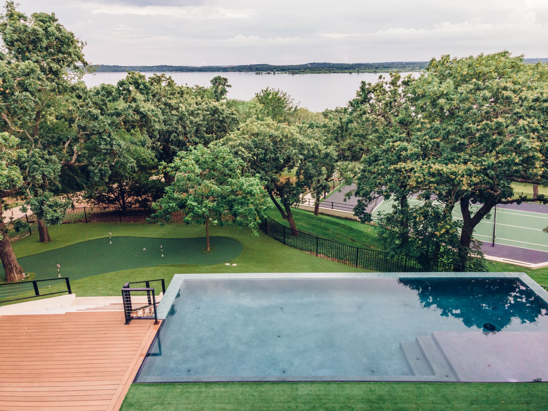 A luxurious backyard with an infinity pool, wooden deck, and well-manicured lawn. Beyond the pool, there's a lawn with a few golf flags, several tall trees, and a tennis court. In the background, a large lake stretches to the horizon under a cloudy sky.