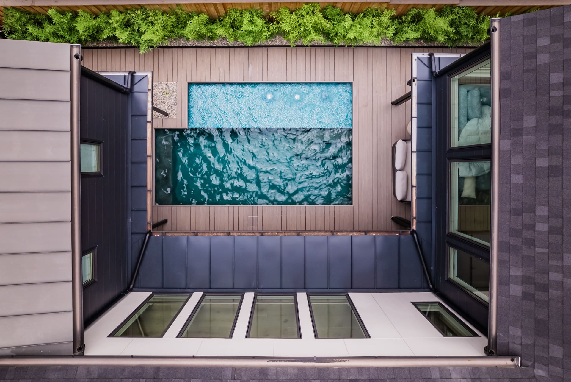 A top-down view of a modern, narrow backyard featuring a rectangular swimming pool with a wooden deck. Two lounge chairs are placed near the pool, and the area is surrounded by lush green plants and two adjacent buildings.