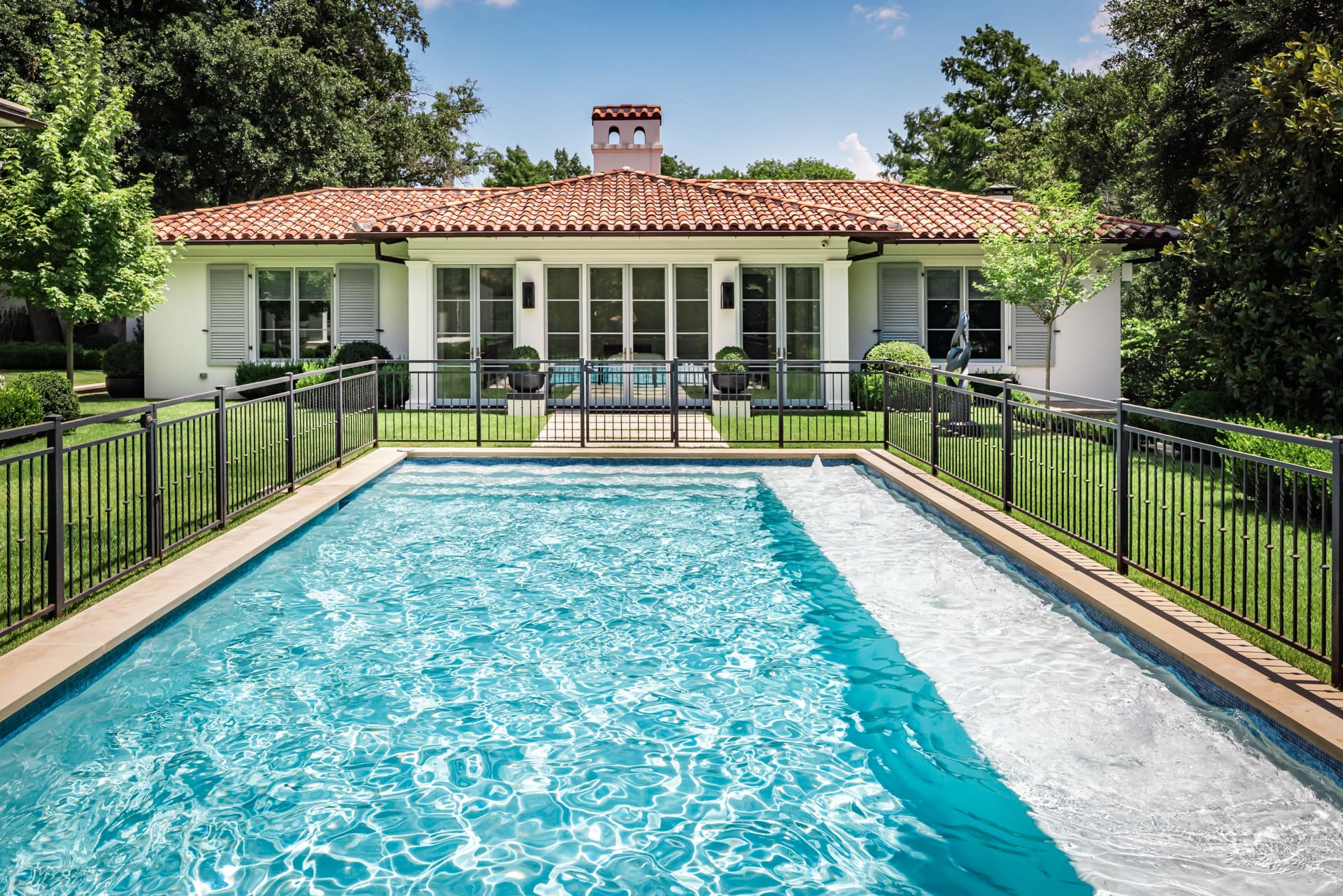 A backyard features a swimming pool with clear blue water, surrounded by a black metal fence. In the background, there is a white house with a red-tiled roof, large windows, and doors. The yard is landscaped with greenery and trees. The sky is clear and blue.