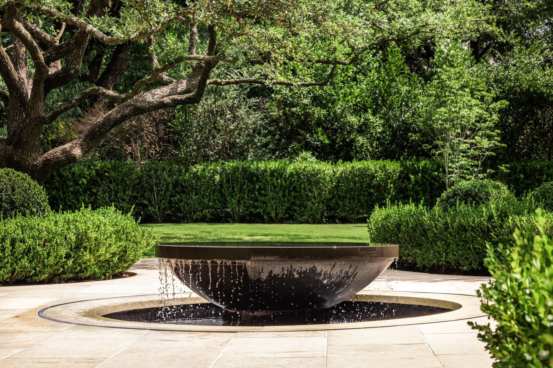 A modern, circular fountain surrounded by lush greenery and hedges. Water cascades from the edges of a central basin into a lower circular pool. Trees with sprawling branches and a well-manicured lawn are visible in the background.