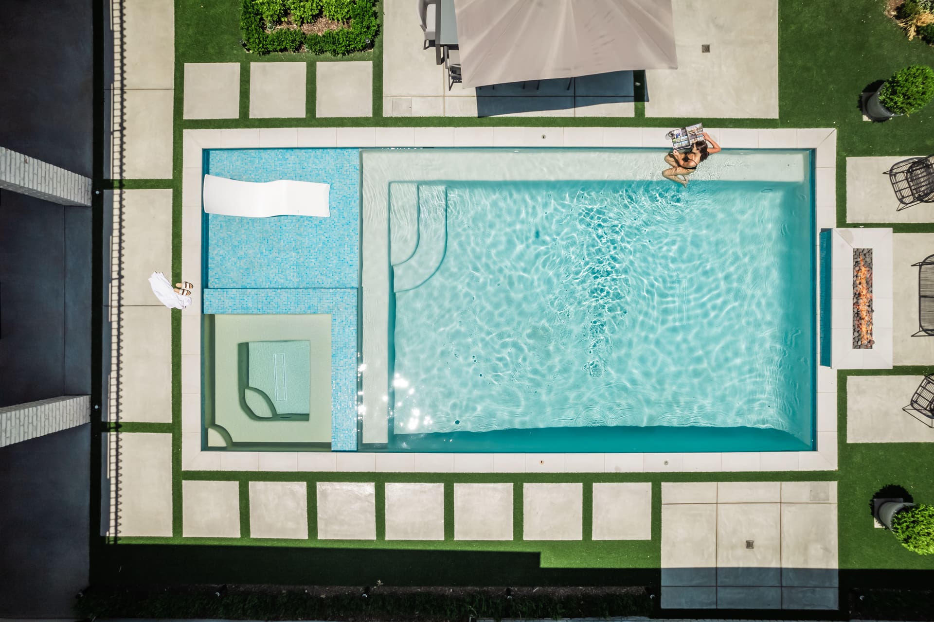 Aerial view of a modern backyard swimming pool area with geometric design. The pool includes a shallow lounge area, adjacent hot tub, and seating space. A person lounges on a float in the water. Poolside features include lounge chairs, a canopy, and greenery.