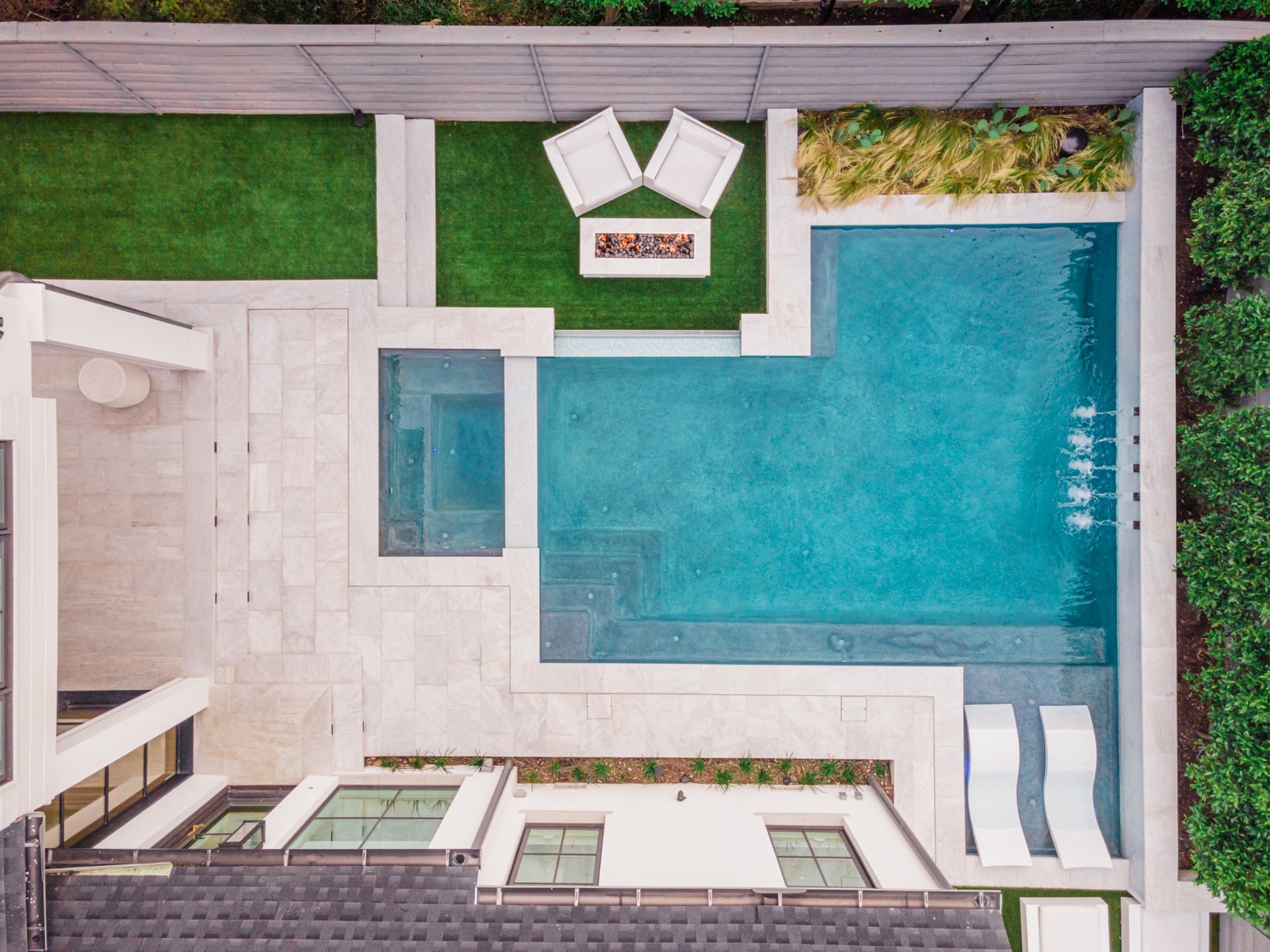 Aerial view of a modern backyard featuring a large rectangular swimming pool with adjacent spa, surrounded by a stone patio. On the lush green lawn, two white outdoor chairs are positioned near a firepit. A fence and greenery provide privacy around the area.