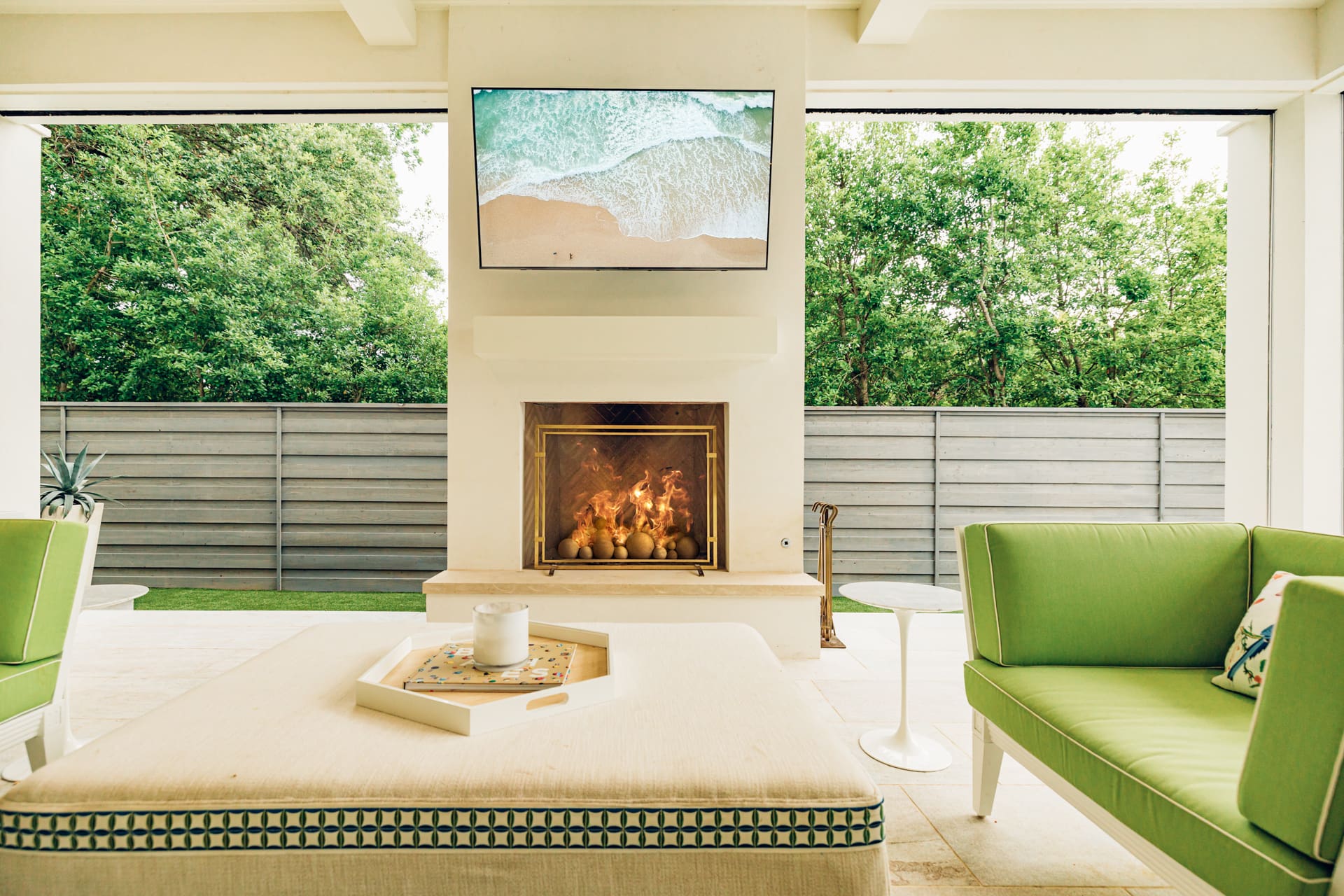 Outdoor living space featuring a cozy fireplace with a fire burning, a wall-mounted TV above showing a beach scene, cushioned green seating, and an ottoman with a tray and candle. The background shows a fence with tall green trees beyond it.