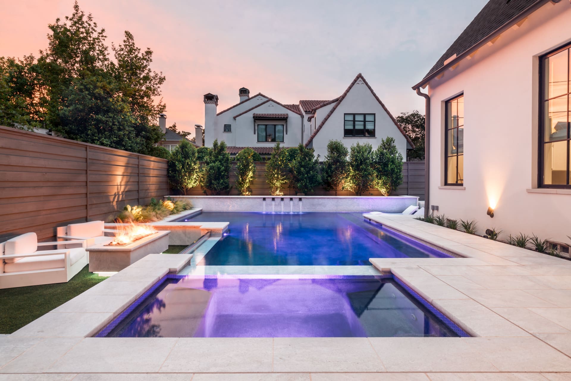 A modern backyard features a swimming pool with purple lighting, a hot tub, and a fire pit area with seating. The space is surrounded by a wooden privacy fence and lush greenery. In the background, two houses with steeply pitched roofs are visible.