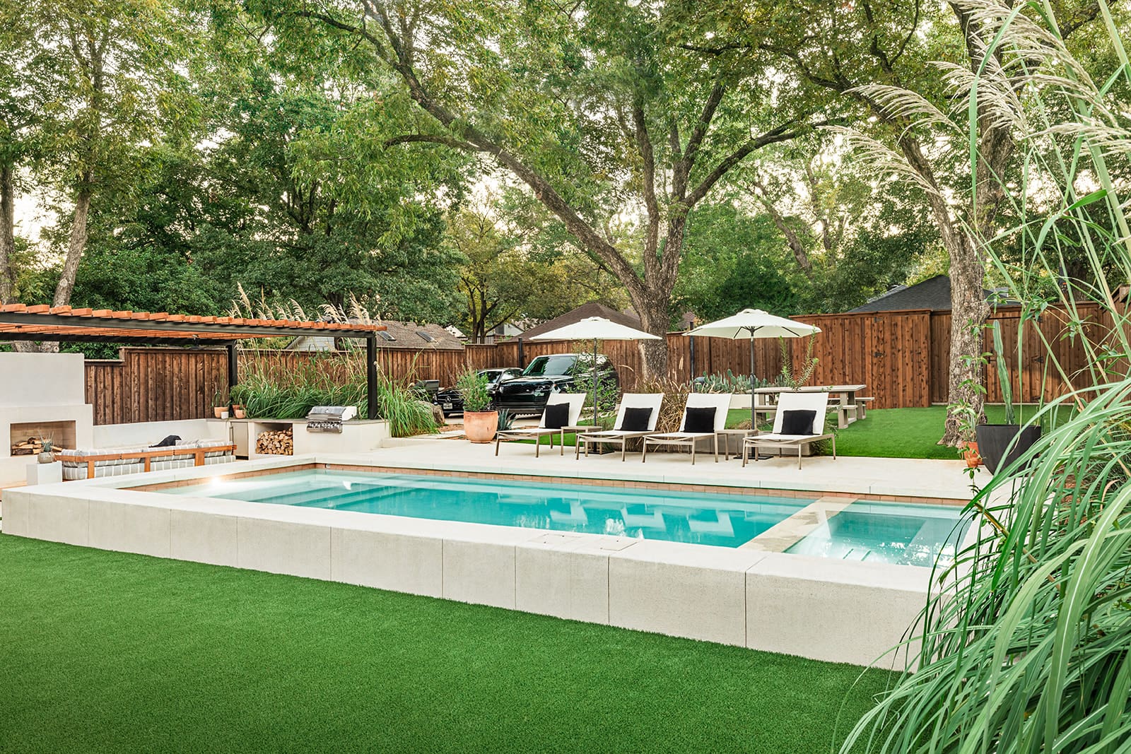 A well-maintained backyard features a rectangular swimming pool surrounded by a neatly trimmed lawn. There are lounge chairs under umbrellas, a covered seating area, and a barbecue grill near a wooden fence. Large trees provide shade in the background.