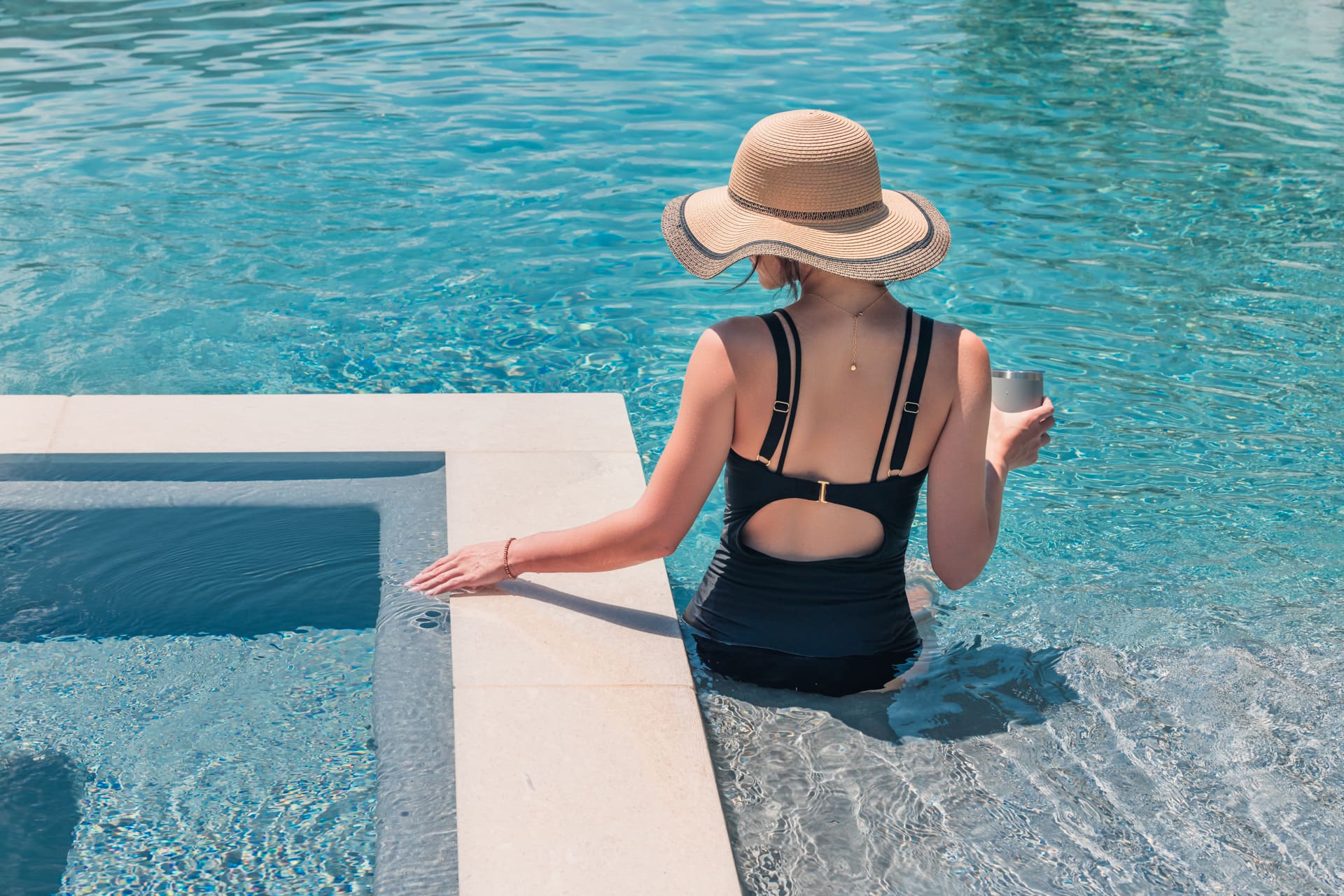 A person wearing a wide-brimmed straw hat and black swimsuit sits on the edge of a pool with one hand on a step and the other holding a drink. They are partially submerged in clear blue water.
