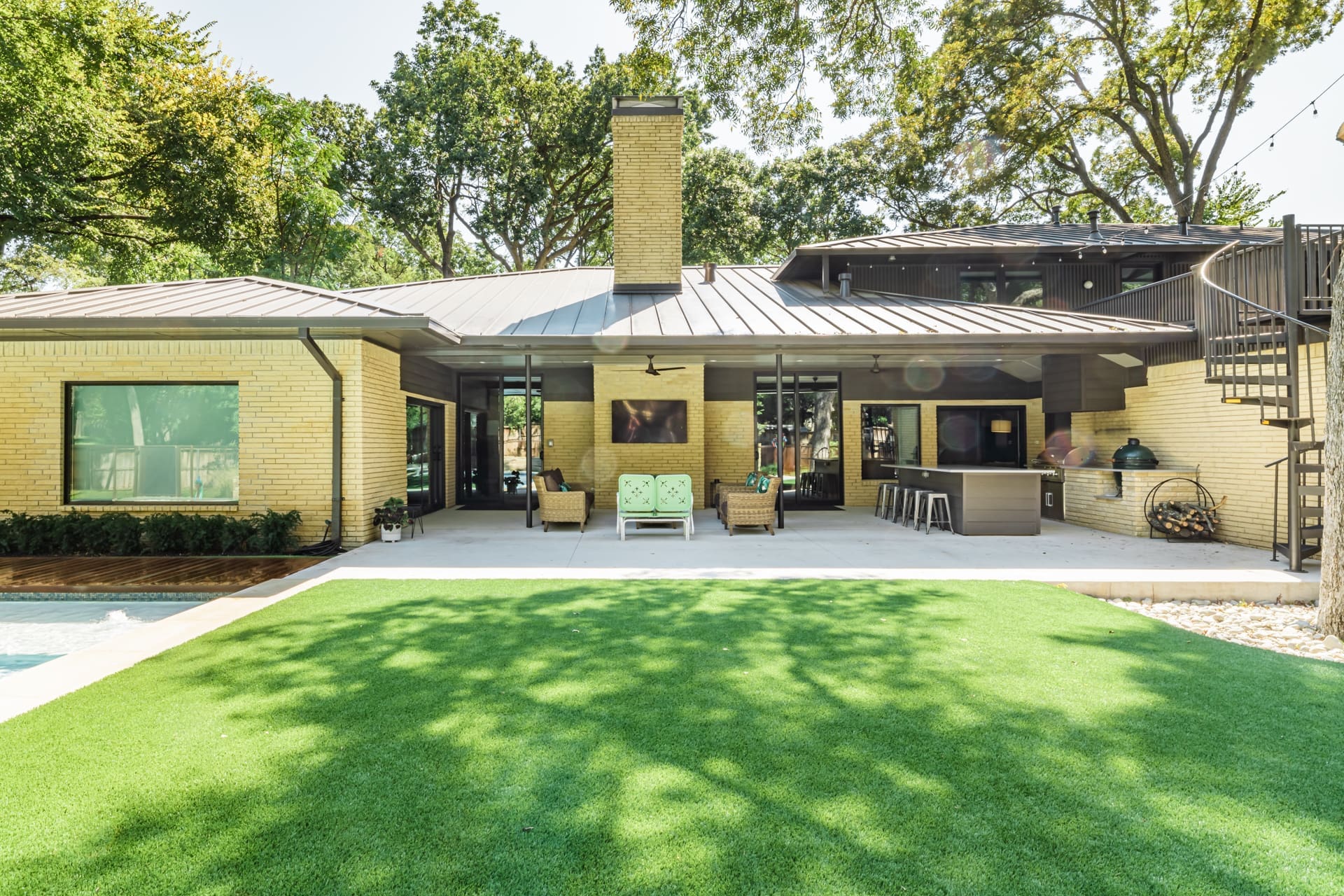 A modern house with a spacious backyard features a large patio area furnished with seating and a dining area. The patio has a roof overhang, and there is an outdoor kitchen. The lawn is well-maintained with a lush green grass, and trees are visible in the background.