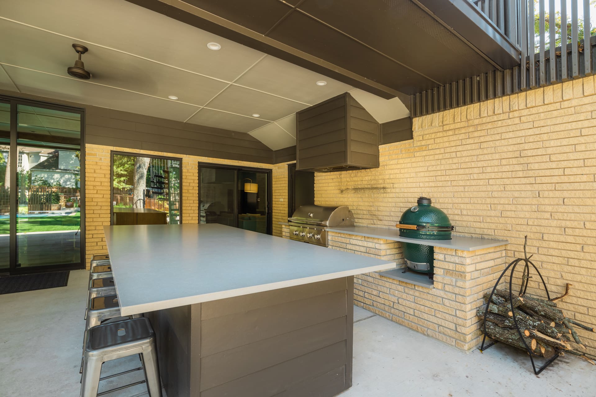 Outdoor kitchen area with a large countertop and bar stools on one side. A built-in grill and a green egg-shaped smoker are situated against a yellow brick wall. A stack of firewood is neatly arranged nearby. Ceiling fan and modern lighting overhead, with a view of the garden.