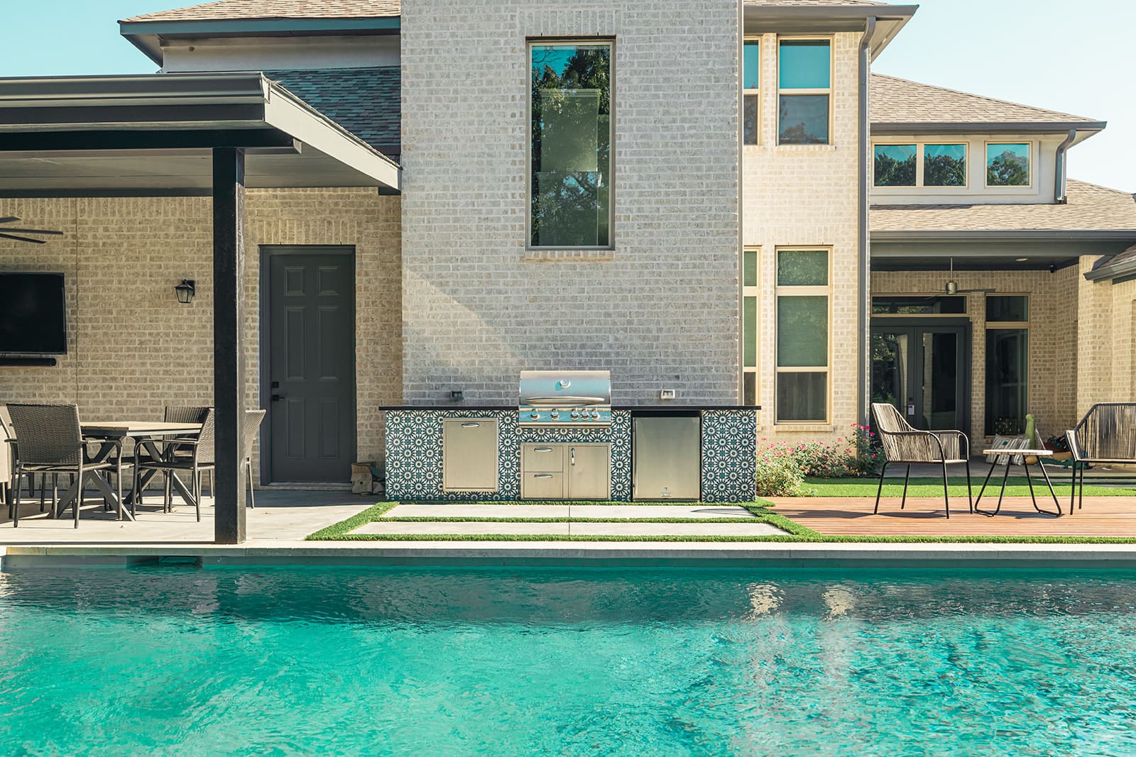 A modern backyard features a pool with turquoise water, an outdoor kitchen with a built-in grill and patterned tile backsplash, a dining area with chairs and a table, and a light-colored brick house in the background with large windows and an upper floor.